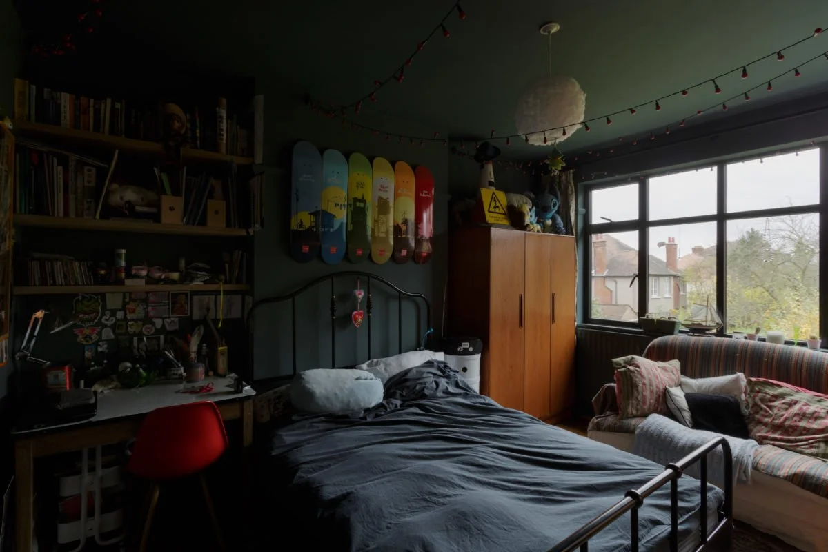 A cozy bedroom with a bed, sofa, bookshelf, skateboard decor, and a large window showing outdoor houses.