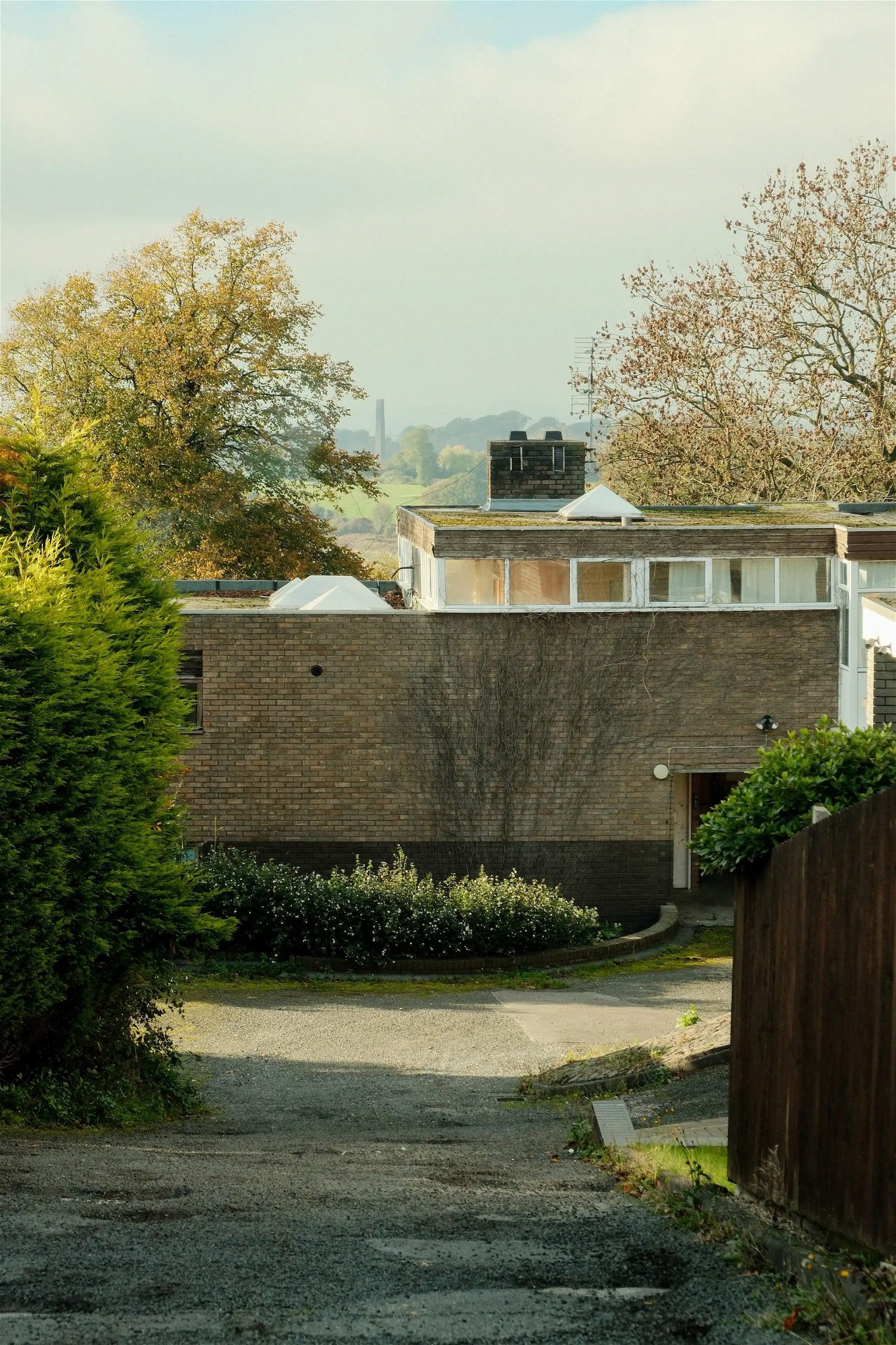 Residential building with trees and plants around, view from a gravel driveway or path in the foreground, amid a suburban or semi-rural setting.