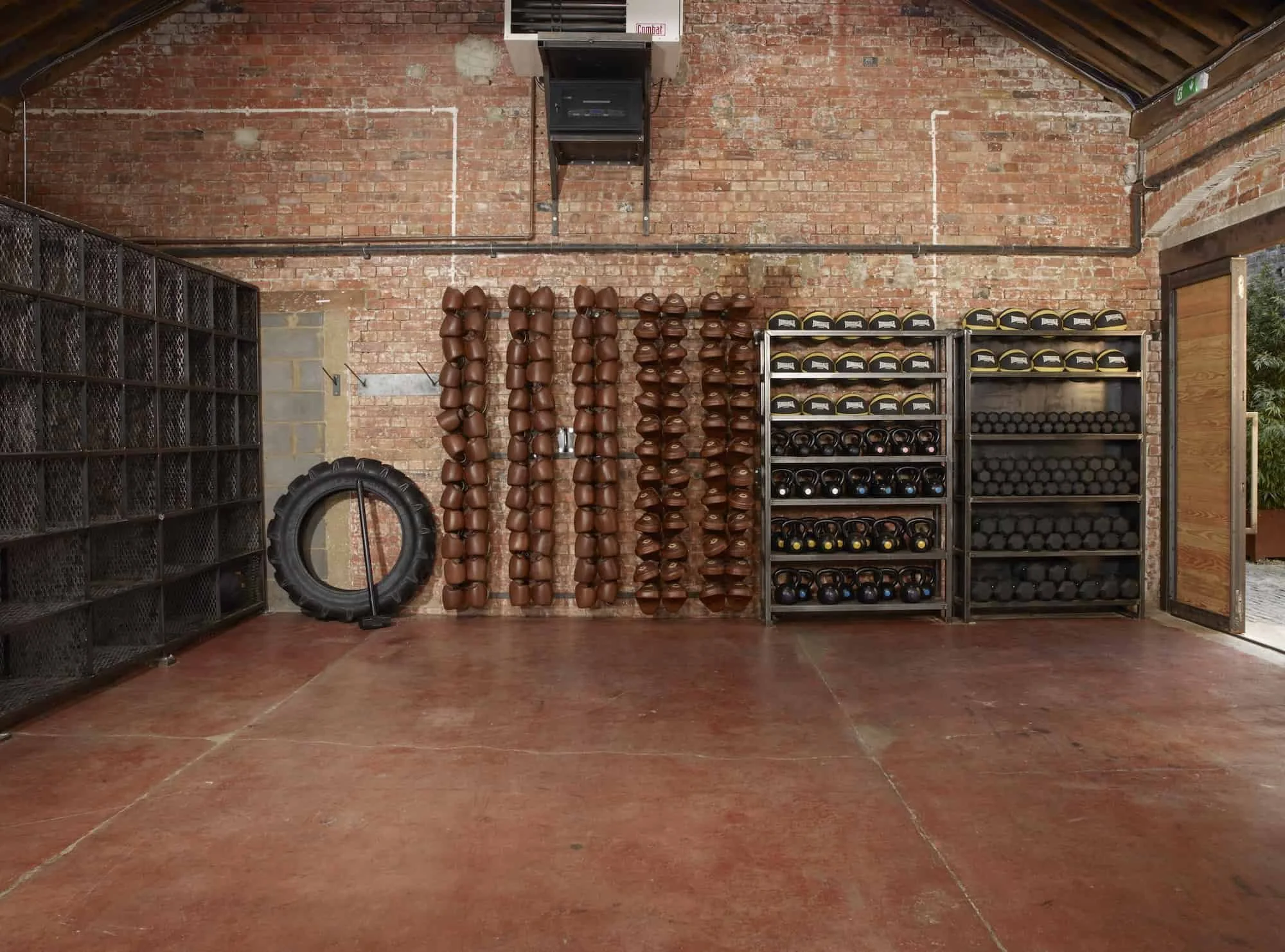 Interior of a gym with brick walls, metal shelving holding black and yellow helmets, kettlebells, and black racks filled with weights, a large tire, and foam rollers or medicine balls on the wall.