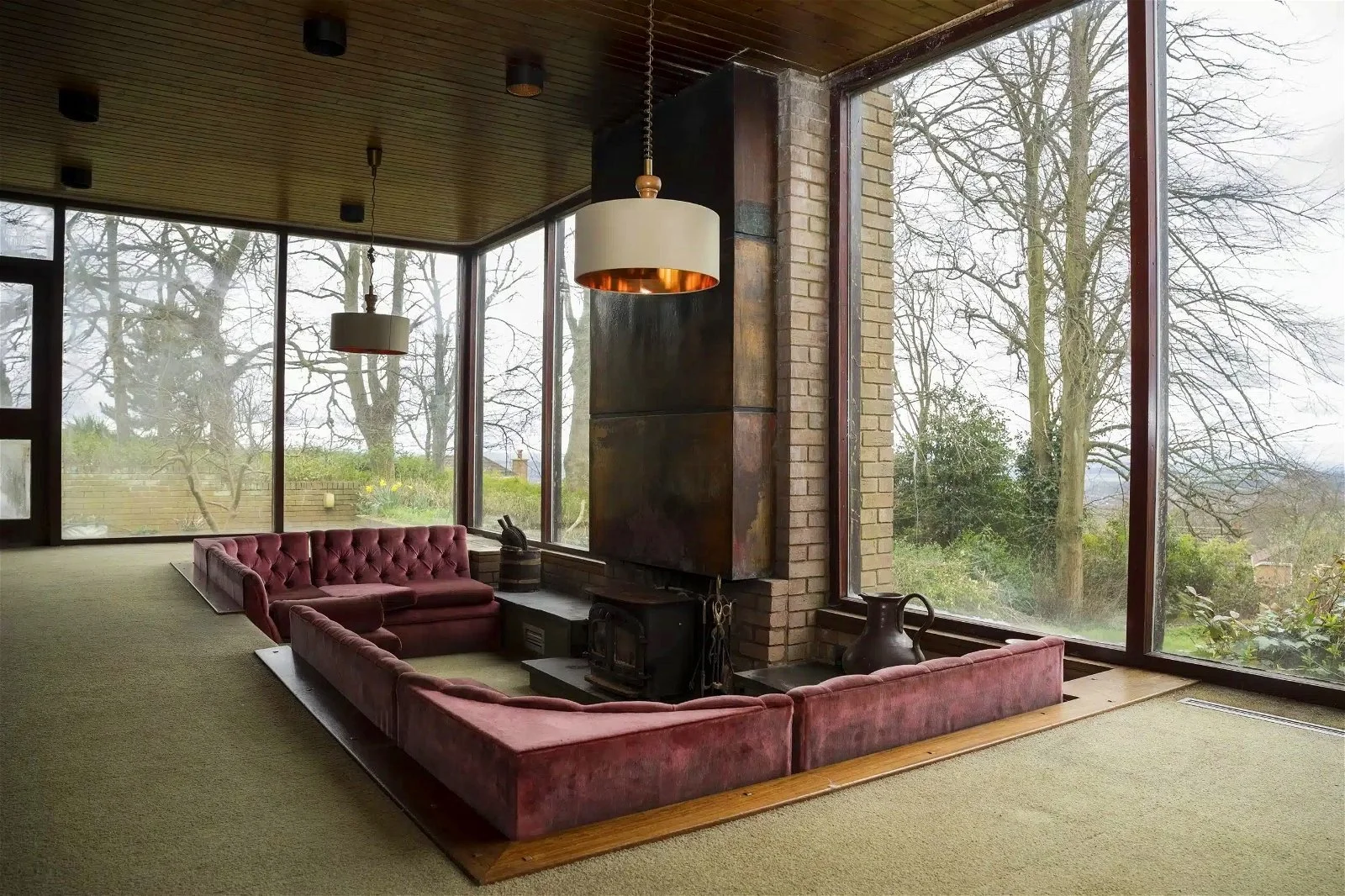 Living room with large floor-to-ceiling windows, a corner sofa with red velvet upholstery, a modern fireplace with a tall metallic chimney, a hanging ceiling light with a white shade, and a view of trees outside.