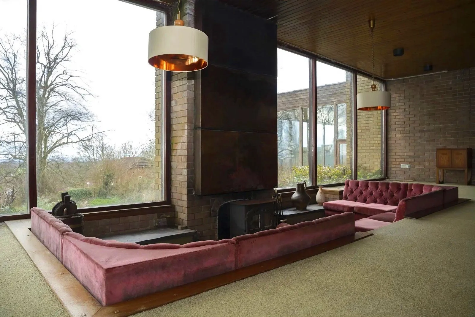 Living room with large floor-to-ceiling windows, brick walls, and a pink velvet sectional sofa arranged around a wood-burning fireplace, with decorative vases on the hearth and modern pendant lights hanging from a wooden ceiling.