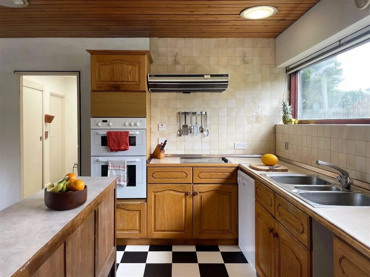 A kitchen with wooden cabinets, a checkered black and white floor, and a large window. There are two bowls of fruit on the counter and a few kitchen utensils hanging on the wall.