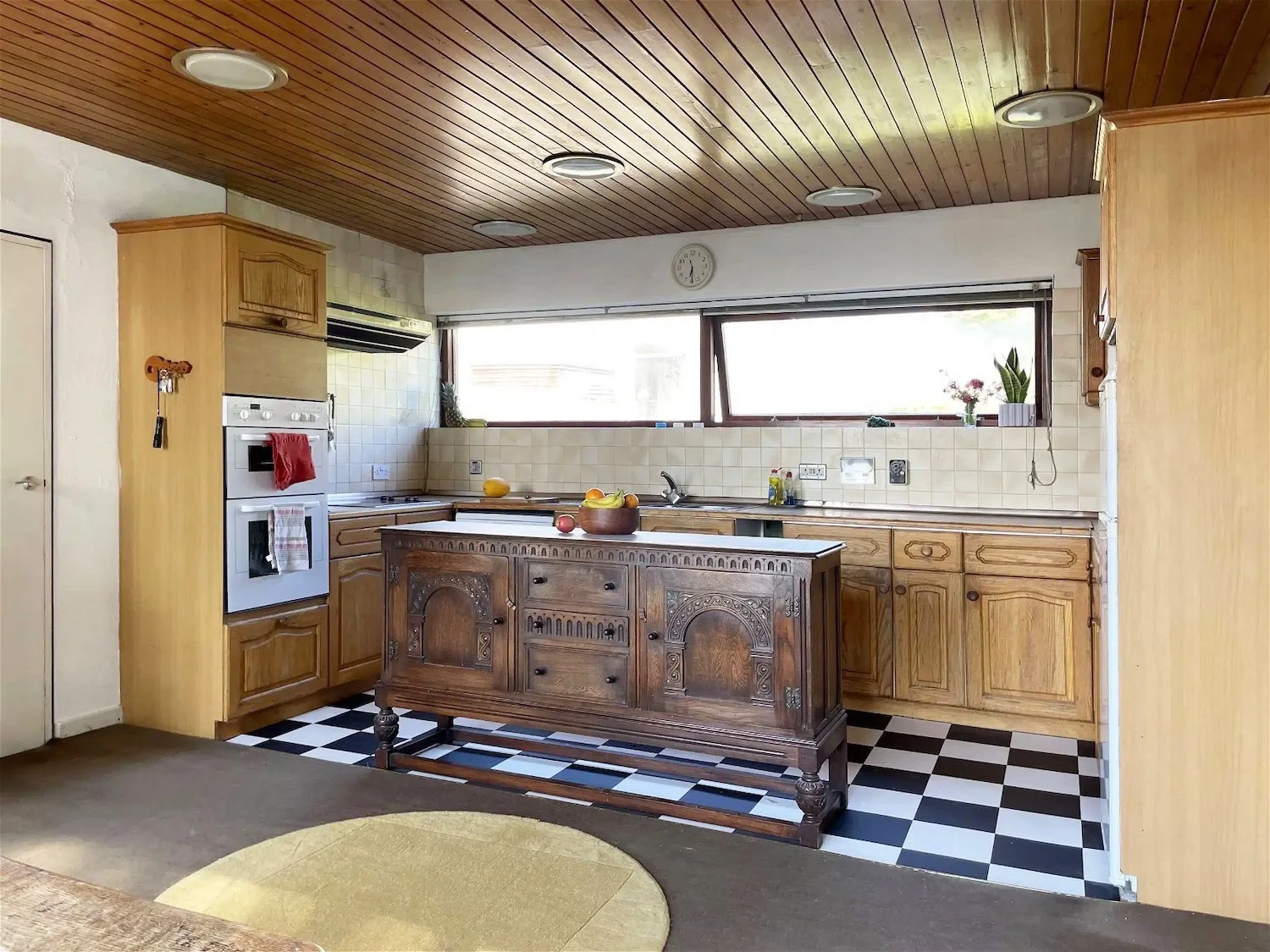 Kitchen with wooden cabinets, a black and white checkered floor, a wooden island, and a long window with plants on the windowsill.