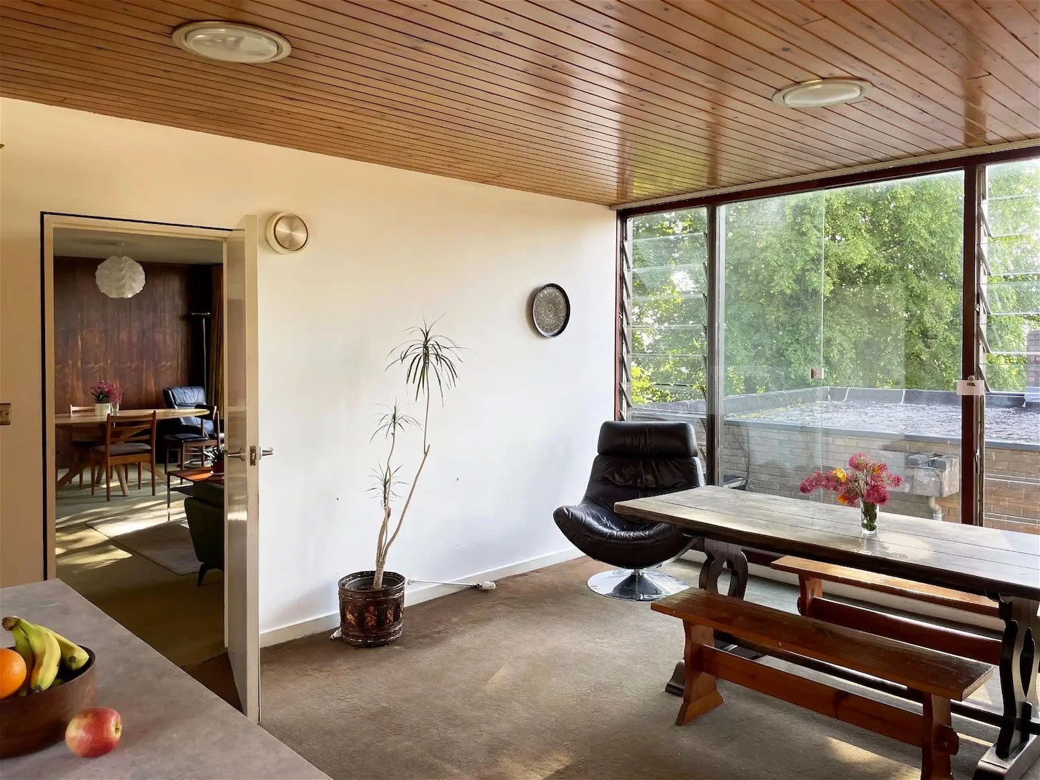 Living room with large glass window, black leather chair, wooden dining table with bench and vase of flowers, potted plant, and view of green trees outside.