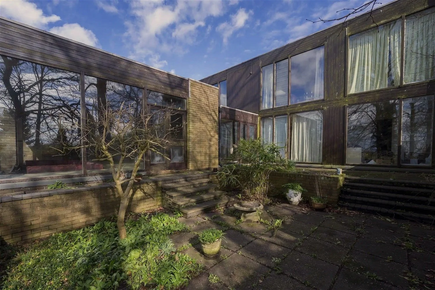 A house with large glass windows and brick exterior, surrounded by a small garden with plants and a leafless tree, under a partly cloudy sky.