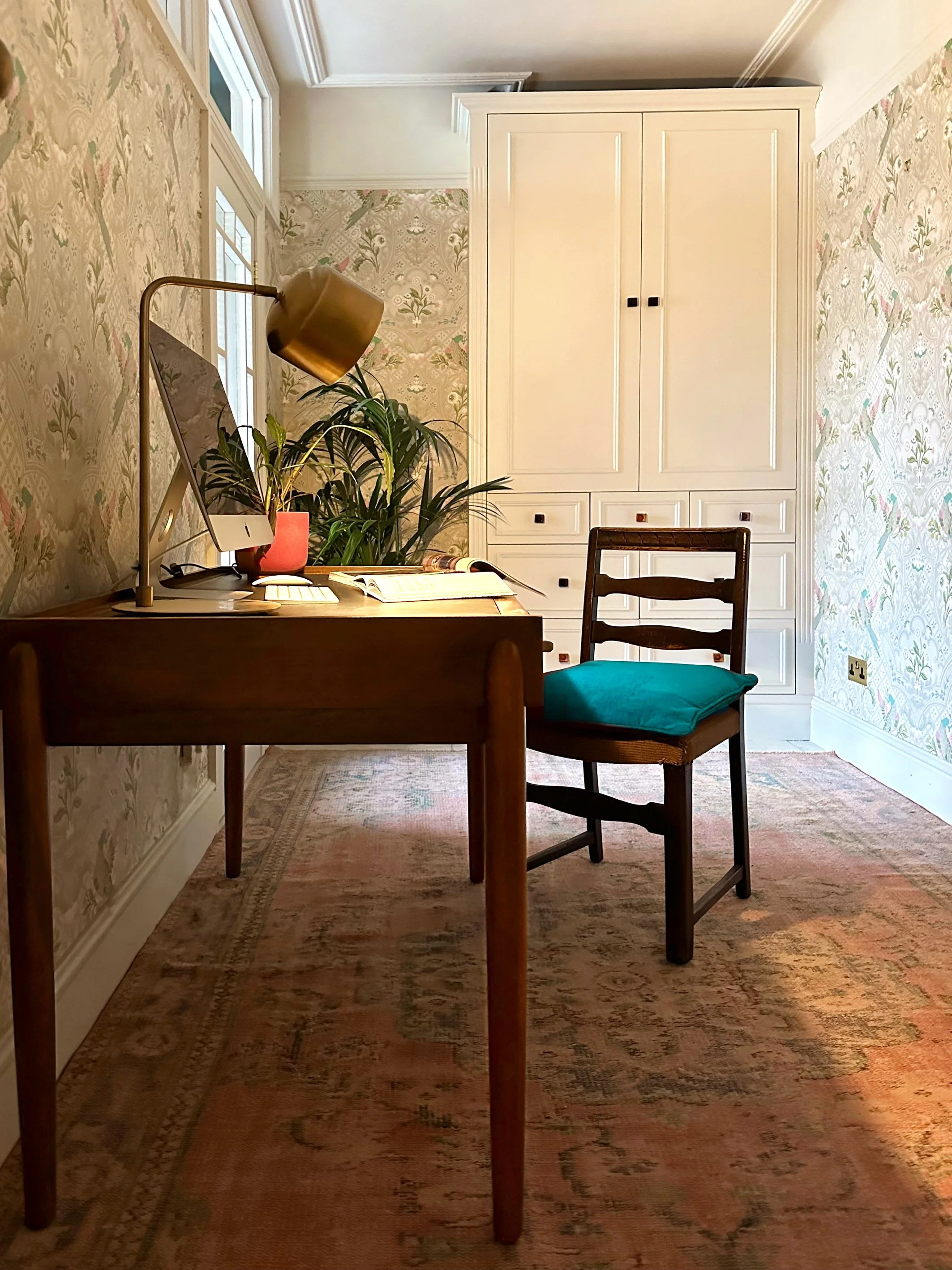 A cozy home office with a wooden desk, an iMac computer, a gold desk lamp, some books, and a large green plant. There is a wooden chair with a teal cushion, a white cabinet, floral wallpaper, and a window with natural light.