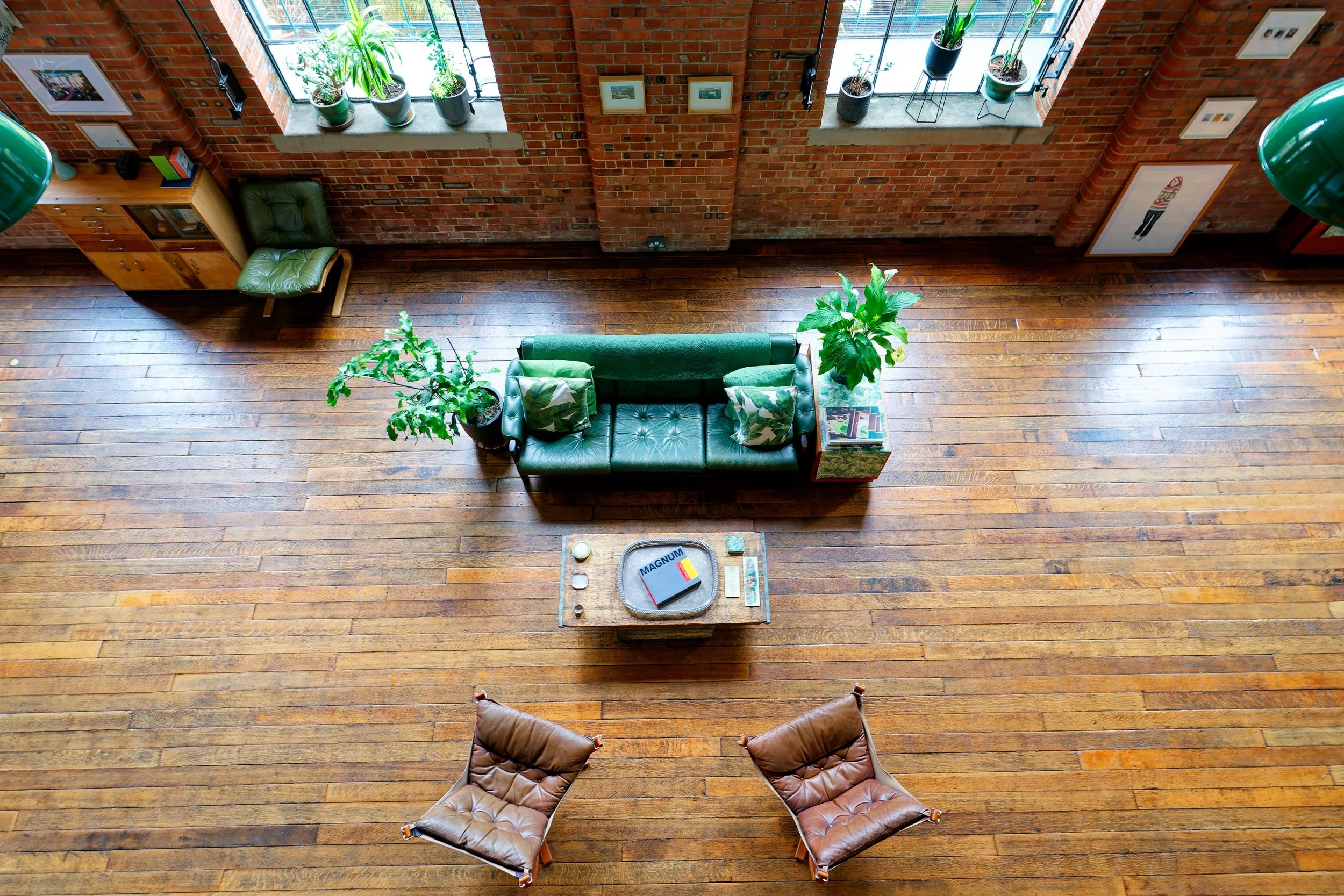 Living room with brick walls, large windows, wooden floor, a green leather sofa, a small wooden coffee table with a tray that says 'Magnum' on it, two brown leather chairs, and several potted plants near the windows.
