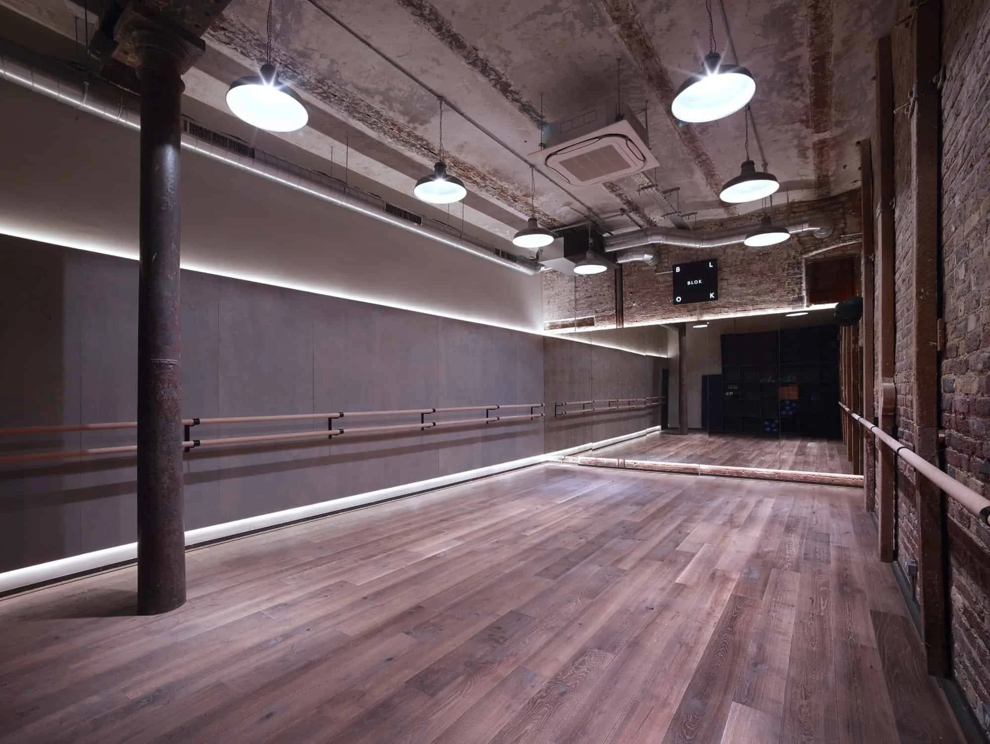 Empty dance studio with wooden flooring, mirrored wall, ballet barres, exposed brick walls, and ceiling-mounted lights.
