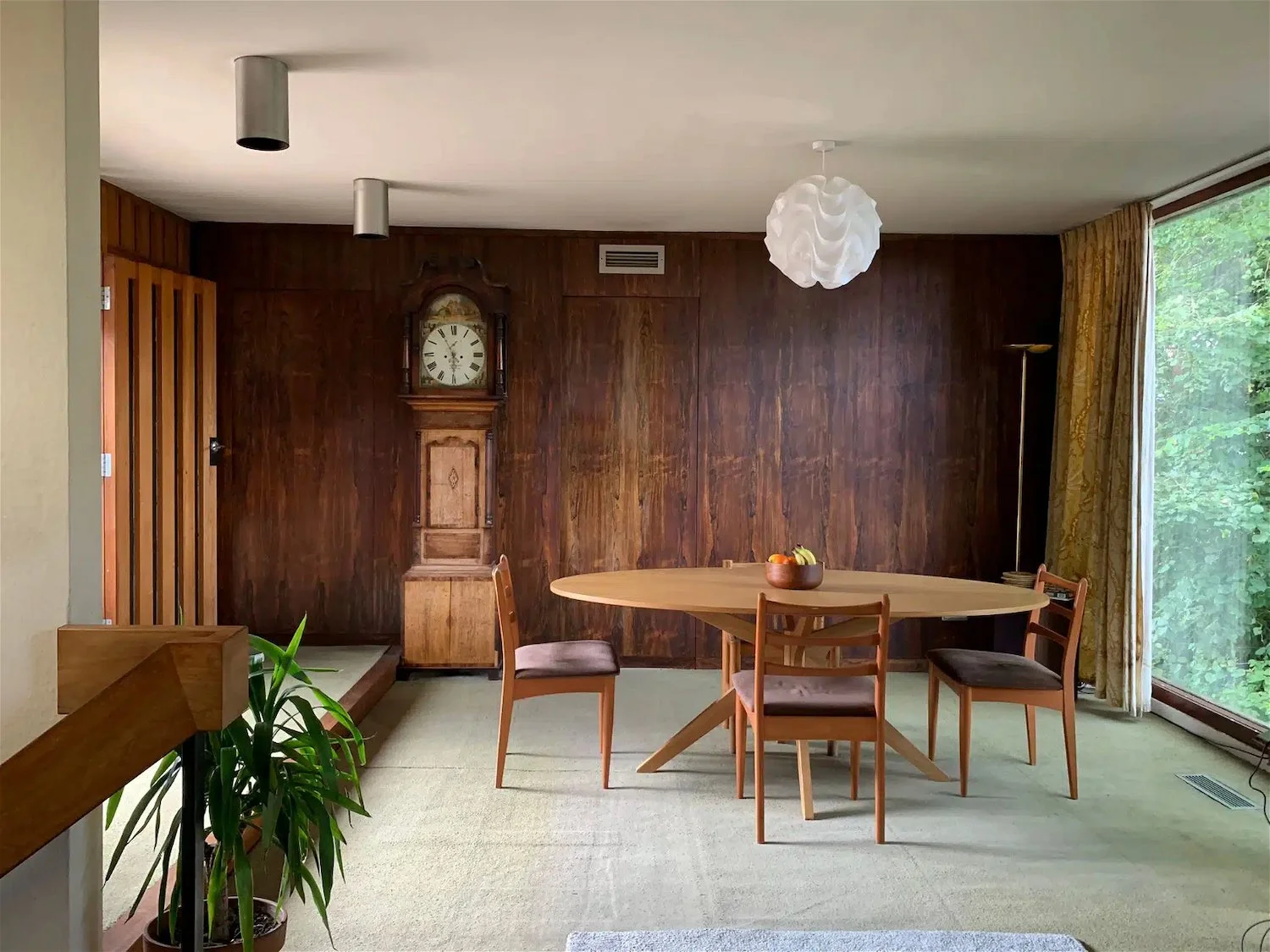 A mid-century dining room with a wooden table and four matching chairs, a vintage wall clock on a wood-paneled wall, a large window with gold curtains showing green foliage, and a white pendant light fixture.