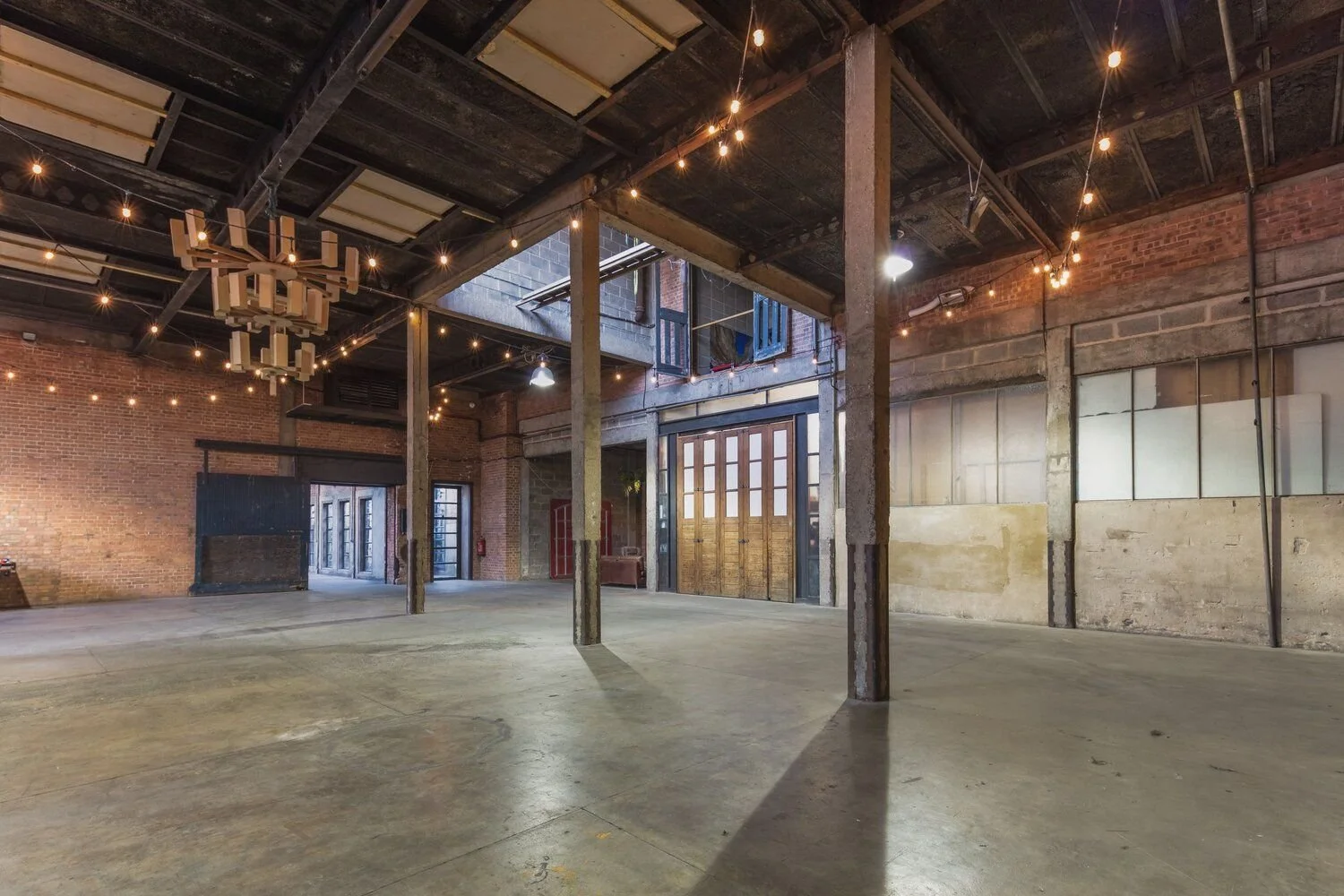 An empty industrial-style event space with brick walls, stained concrete floors, exposed beams, and hanging string lights. The space features a large open area, a loft or second level, and glass doors.