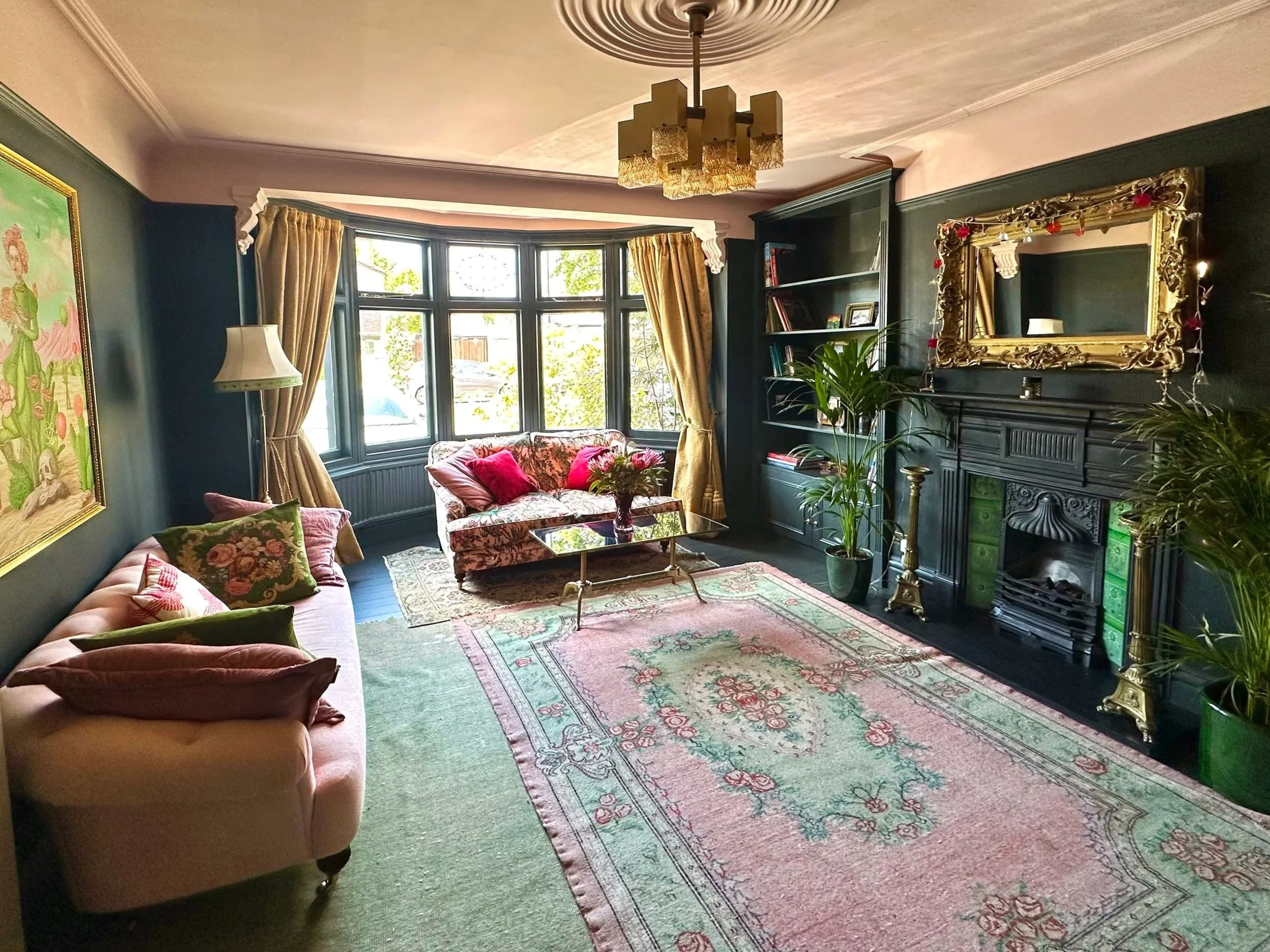 A cozy living room with dark green walls, a large bay window with beige curtains, pink sofas with floral and velvet pillows, a floral patterned area rug, a dark fireplace with ornate gold mirror above, potted plants, and a vintage lamp.