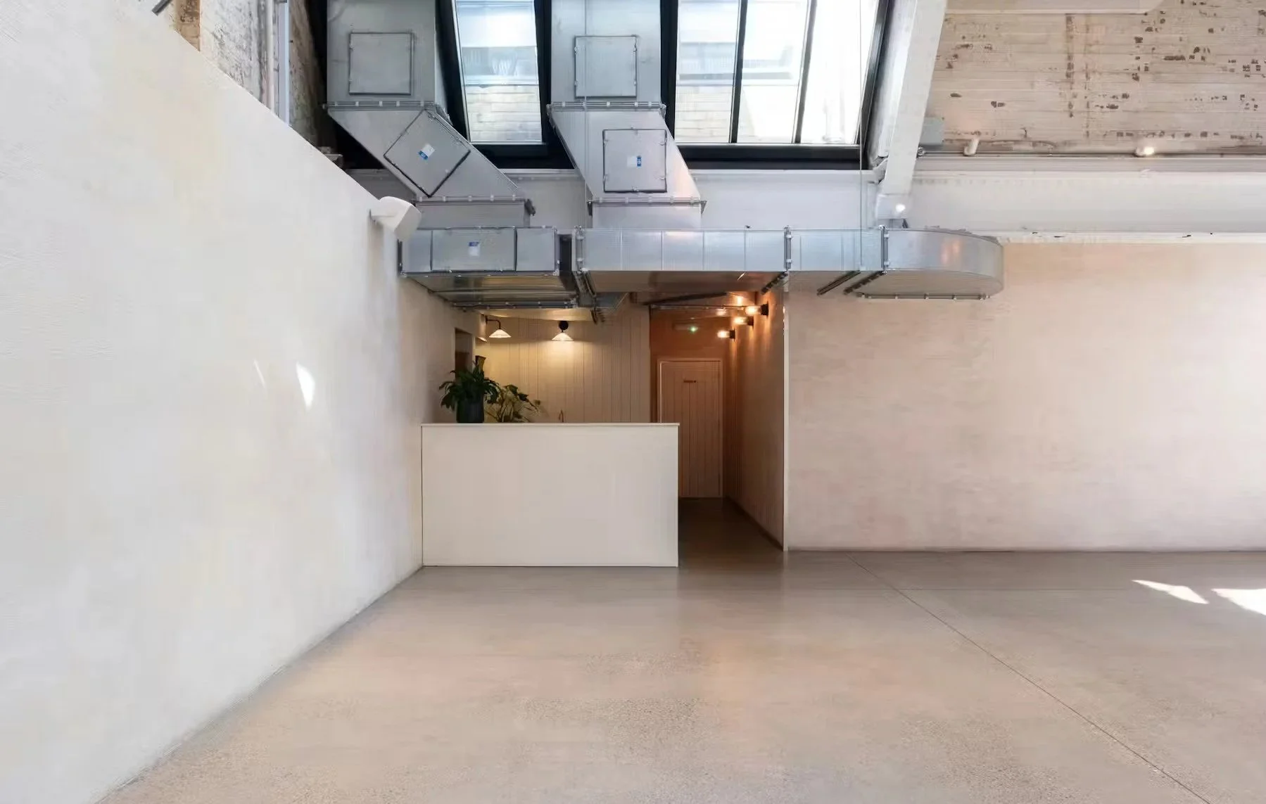 Empty modern interior space with white walls, polished concrete floor, a white reception counter with plants, and an exposed HVAC system with large vents and ducts mounted near the ceiling.