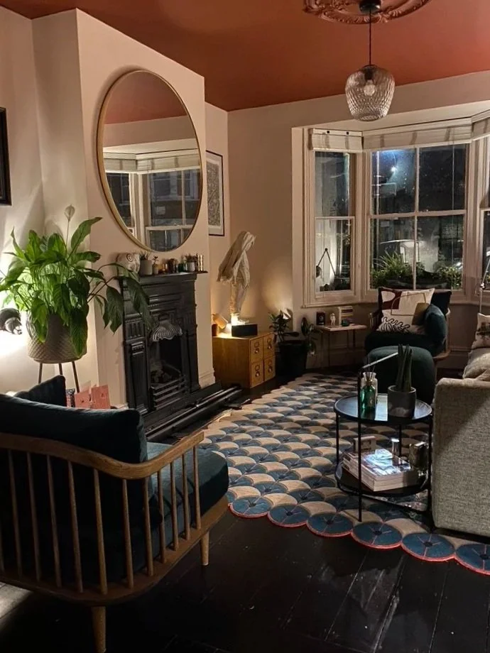 A cozy living room with a fireplace, a large mirror above it, a big window with plants, a patterned rug, and various seating options including a wooden armchair and upholstered chairs, decorated with plants and candles.