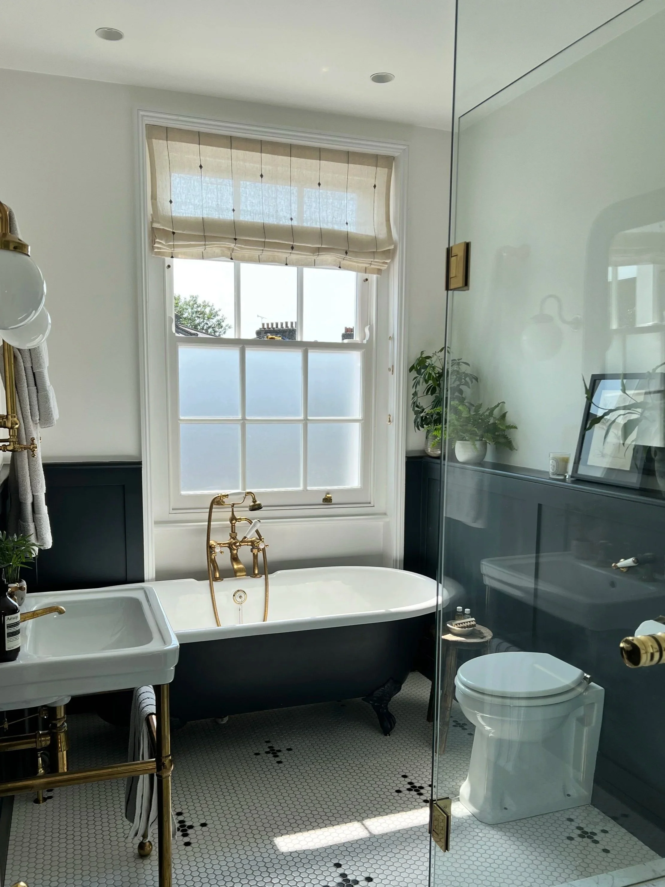 A bathroom with a free-standing bathtub, white tile floor with black accents, a glass shower door, a toilet, a small sink, and plants by the window.