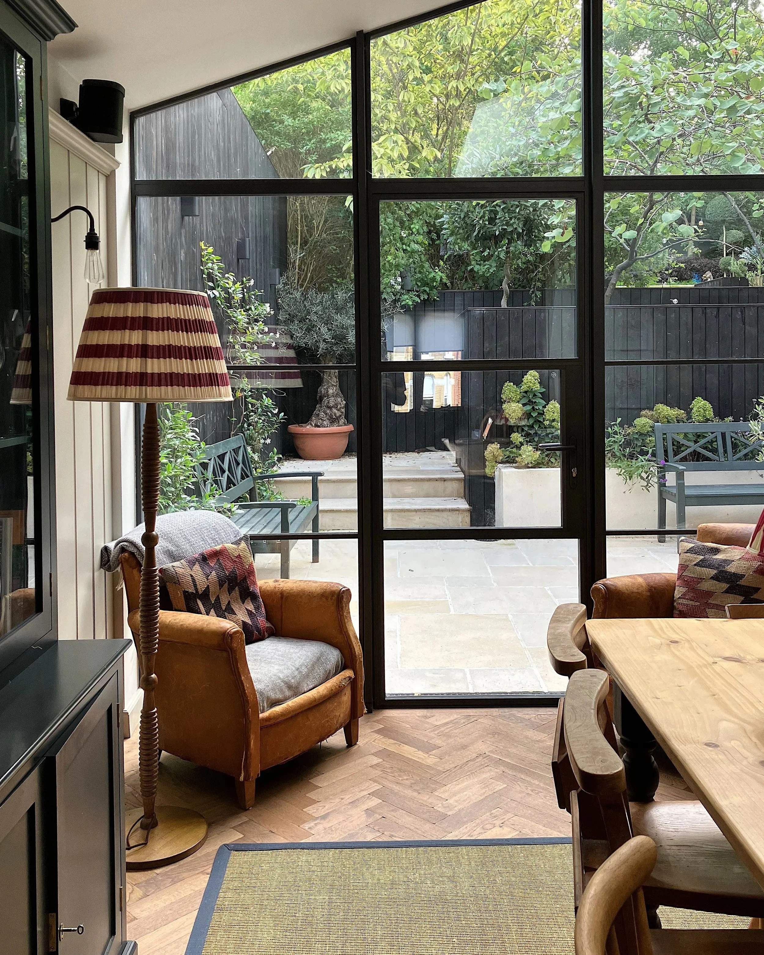 A cozy indoor living space with a view of a backyard garden through large black-framed glass doors. Inside, there are armchairs, a floor lamp with a striped red and beige shade, and a wooden dining table with chairs.