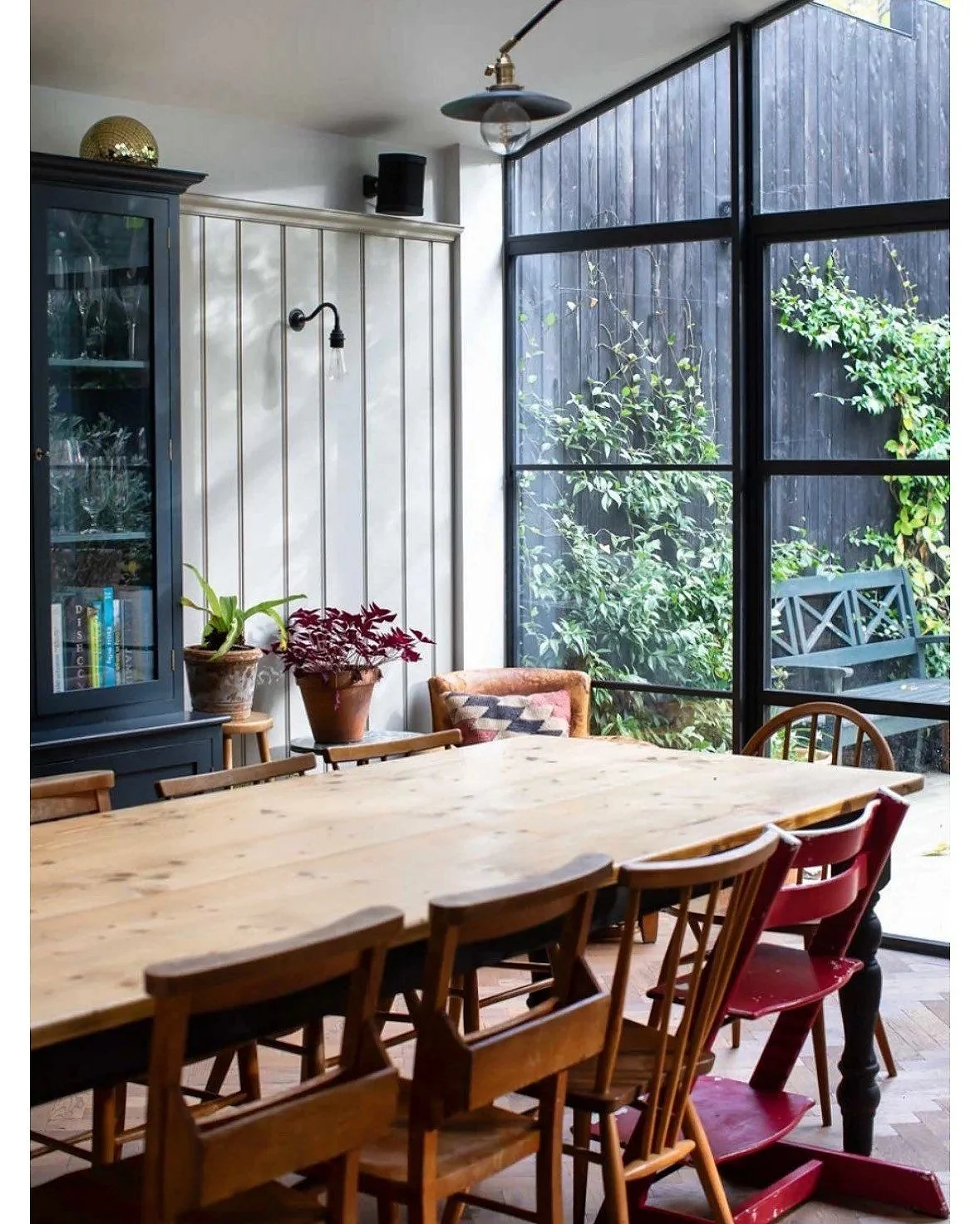A cozy dining area with a large wooden table surrounded by mixed wooden chairs, a black cabinet with glass doors, potted plants, and large windows revealing a garden outside.