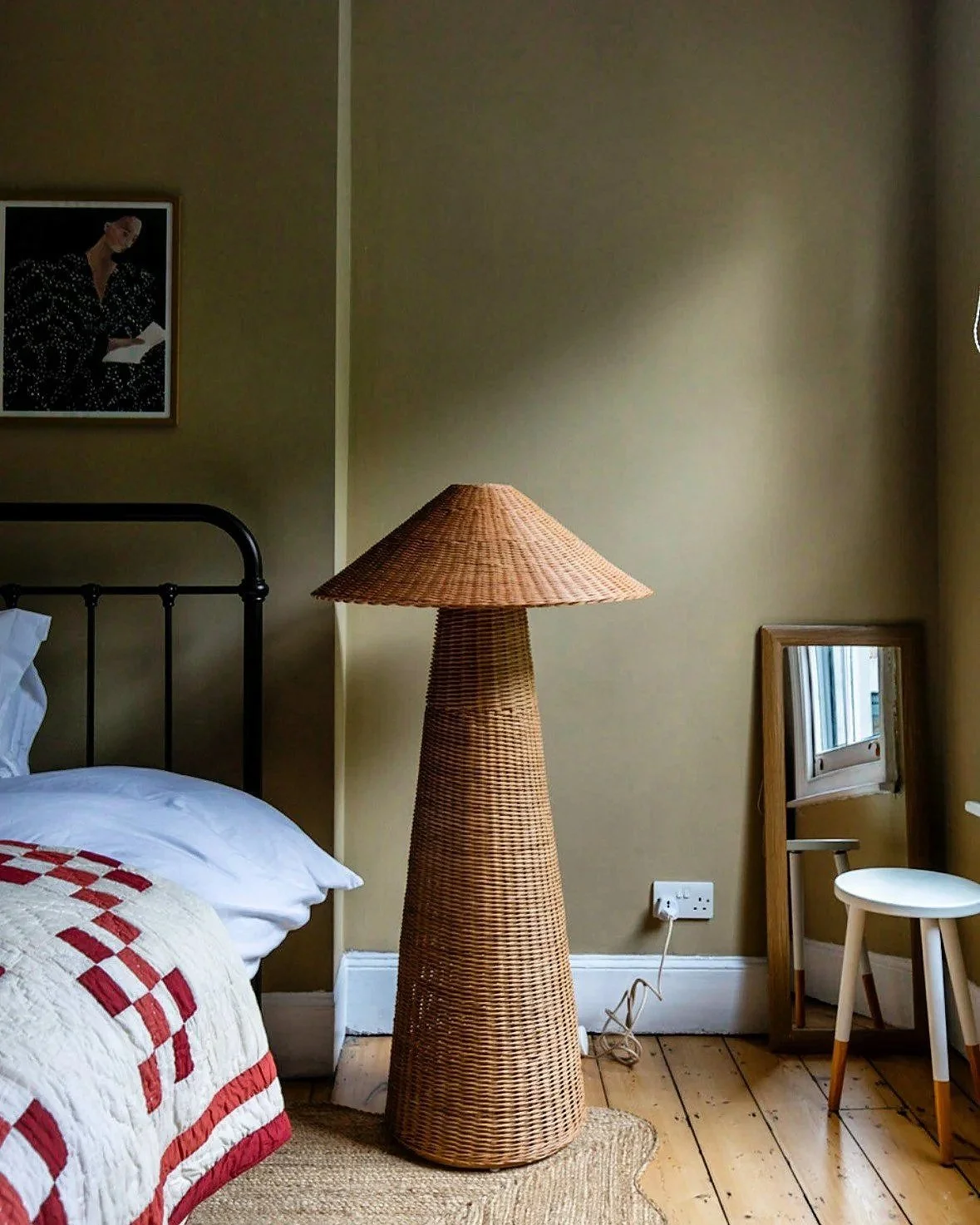 A woven floor lamp shaped like a cone in a bedroom next to a bed with a white pillow and red quilt, a framed picture on the wall, a mirror, a small white stool, and an electrical outlet on the beige wall.