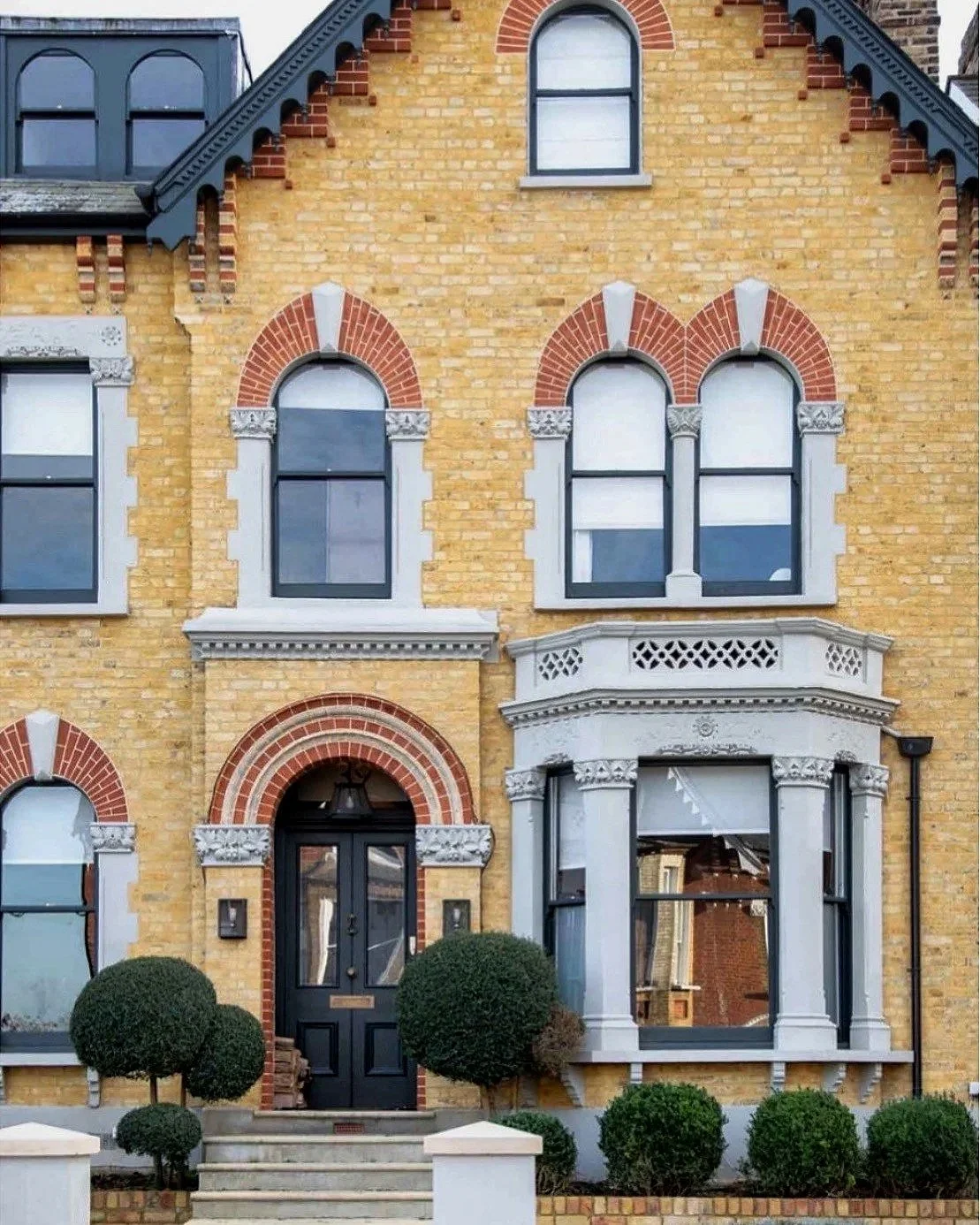 A yellow brick Victorian-style house with arched windows framed with red brick and decorative white trim. The house has a black front door with a semi-circular brick arch, a small front porch with steps, and neatly trimmed bushes in front.