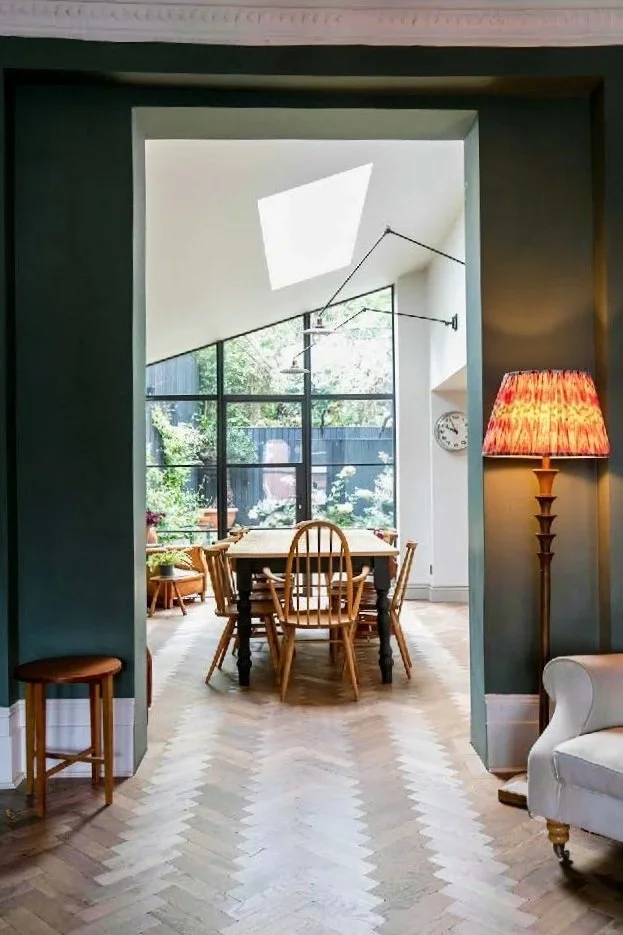View through an archway showing a dining room with a wooden table and chairs, large window with greenery outside, and a skylight ceiling, with a floor lamp and a clock on the wall.