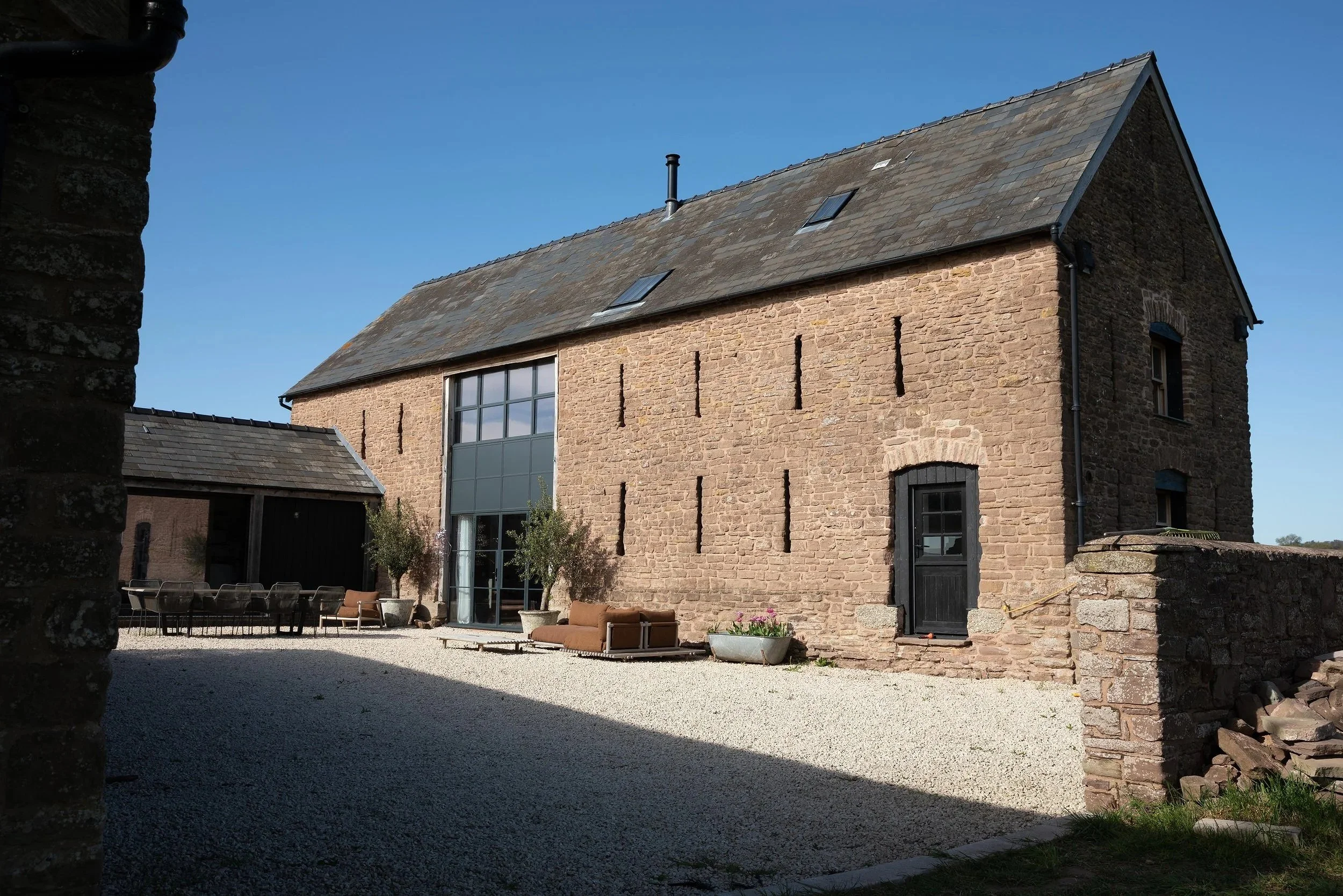 A rustic stone house with a sloped roof, large windows, and a modern glass wall section, set against a clear blue sky. There are outdoor seating and potted plants outside.