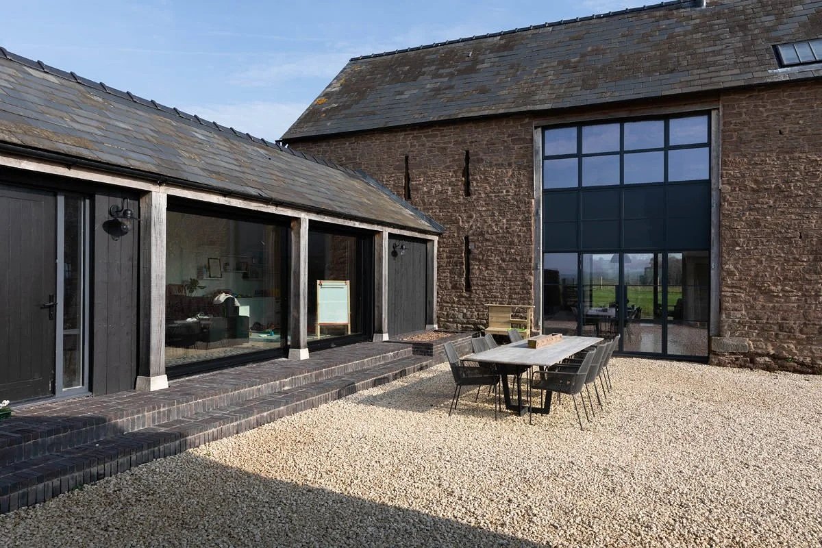 Outdoor patio with a dining table and chairs in front of a rustic brick building with large glass windows and black wooden accents.