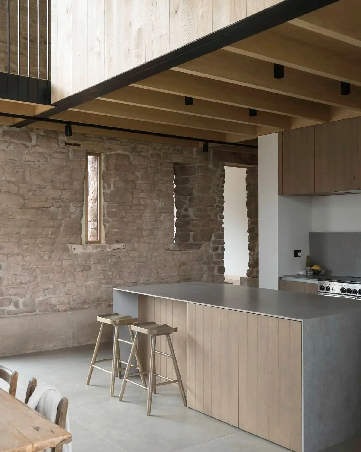 Modern kitchen with stone walls and wooden accents, featuring a concrete countertop island with two wooden stools, and an open doorway leading to another room.