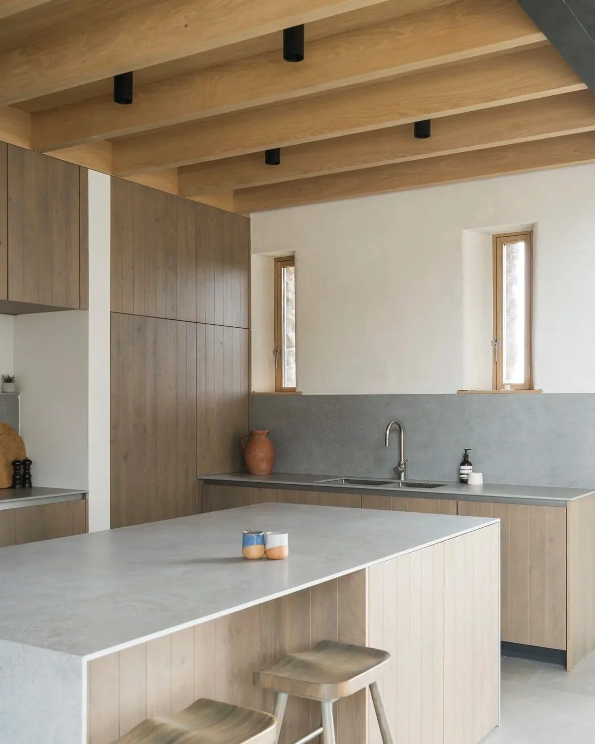 Minimalist kitchen with wooden cabinetry, a large white island, two small candles on the island, a terracotta vase above the sink, and small windows letting in natural light.