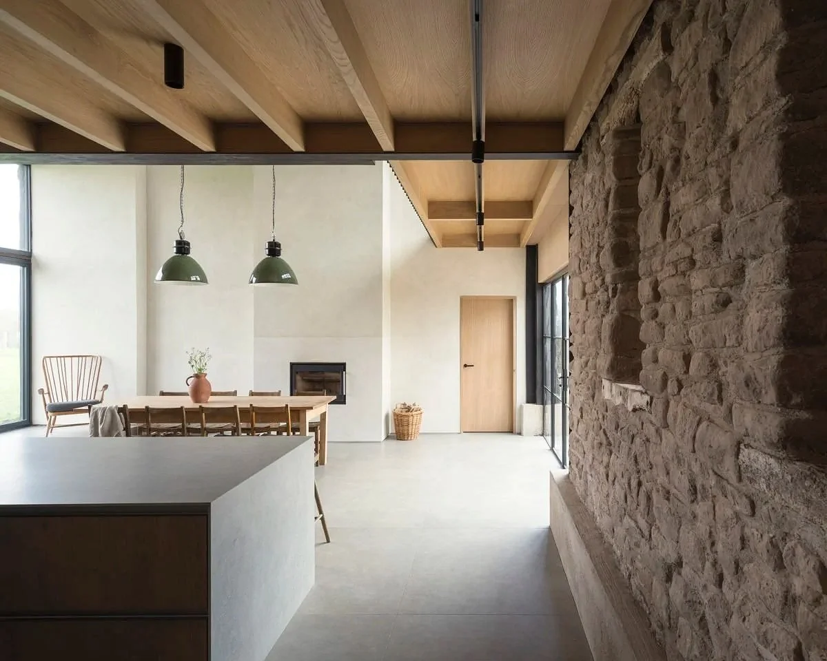 Modern open-concept living and dining area with a stone accent wall, wooden ceiling beams, large windows, and minimalist furniture.
