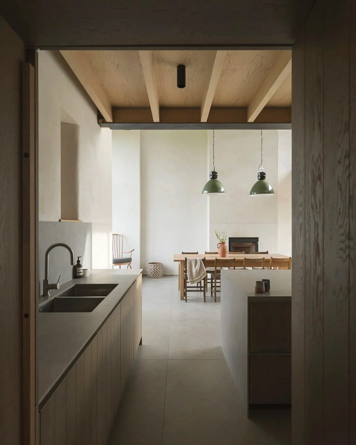 View through a doorway into a minimalist kitchen and dining area with a wooden table, green pendant lights, and a simple potted plant on the table.
