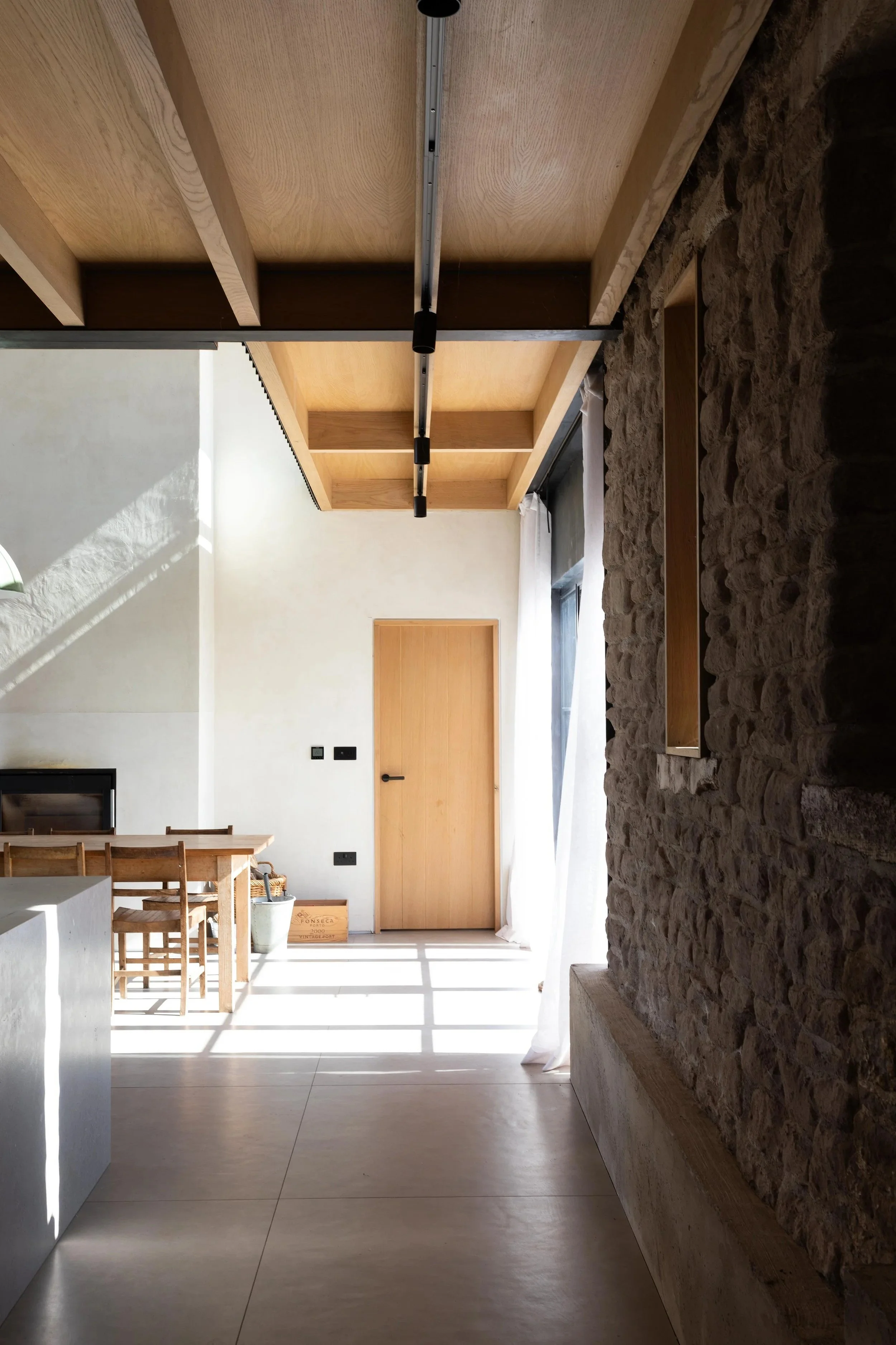 Interior view of a modern room with a high ceiling, wooden beams, a stone wall on the right, a wooden door, a window with sheer white curtains, and a small dining table with chairs.