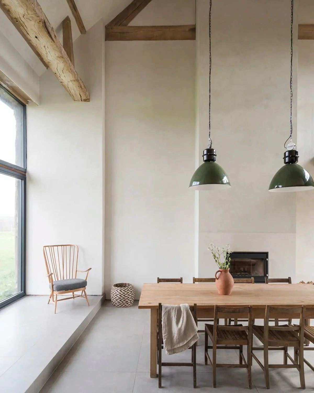 Interior of a minimalistic dining room with a wooden table, six chairs, a vase with flowers, two green pendant lights, a fireplace, a wooden armchair with a cushion, a woven basket, and large windows with natural light.