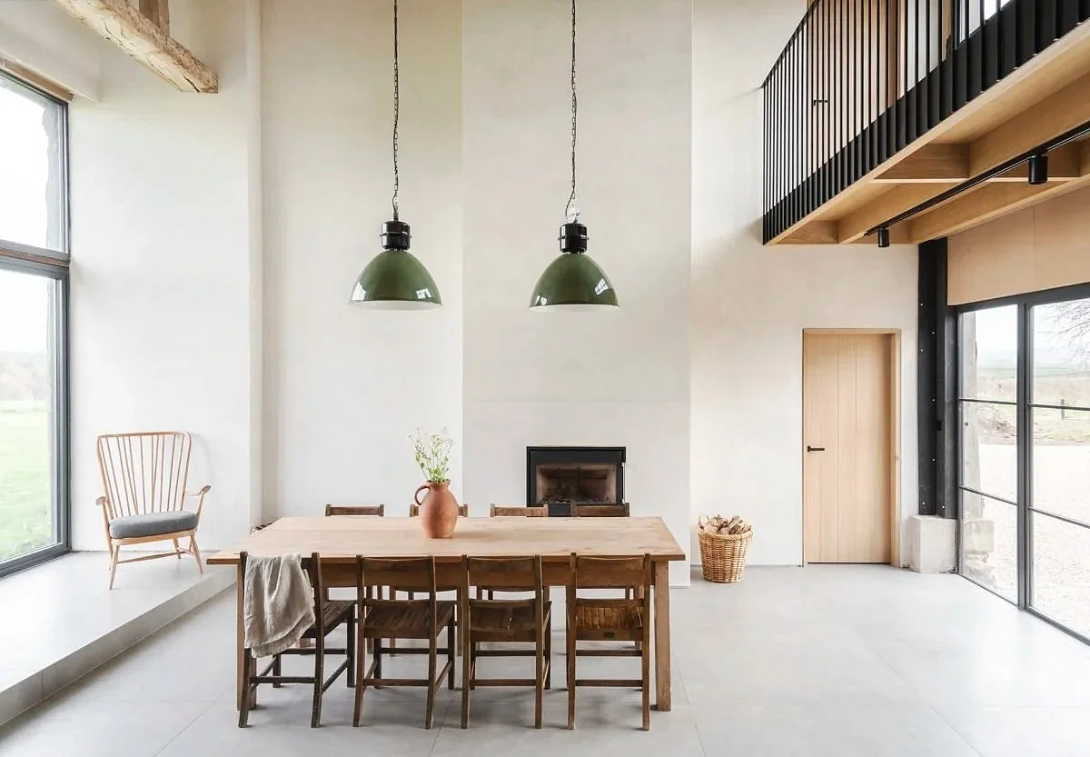 Modern dining area with a wooden table, six chairs, a ceramic vase with flowers, a fireplace, large windows, a wooden door, and a balcony railing in a minimalist, bright room.
