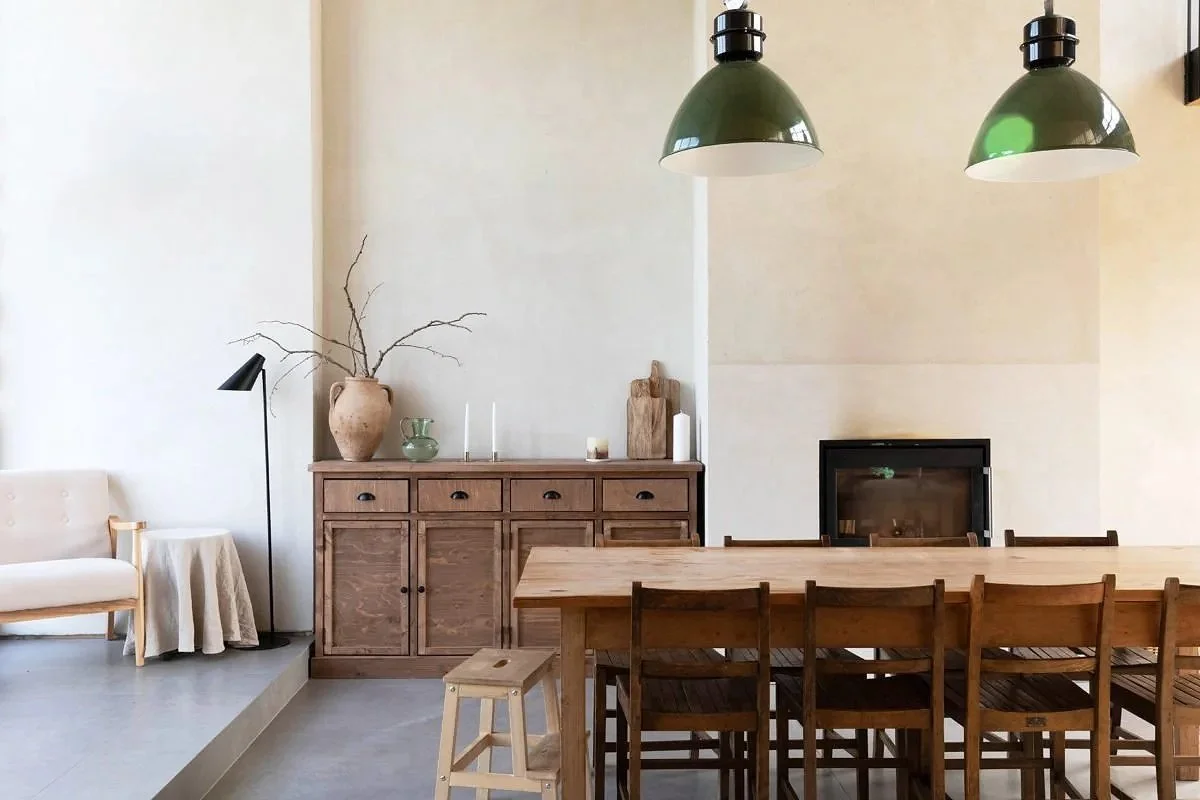 A dining room with a wooden table and chairs, a sideboard with decorations, a bench and a table with a cloth, large lamps hanging from the ceiling, a fireplace, and a minimalist decor style.