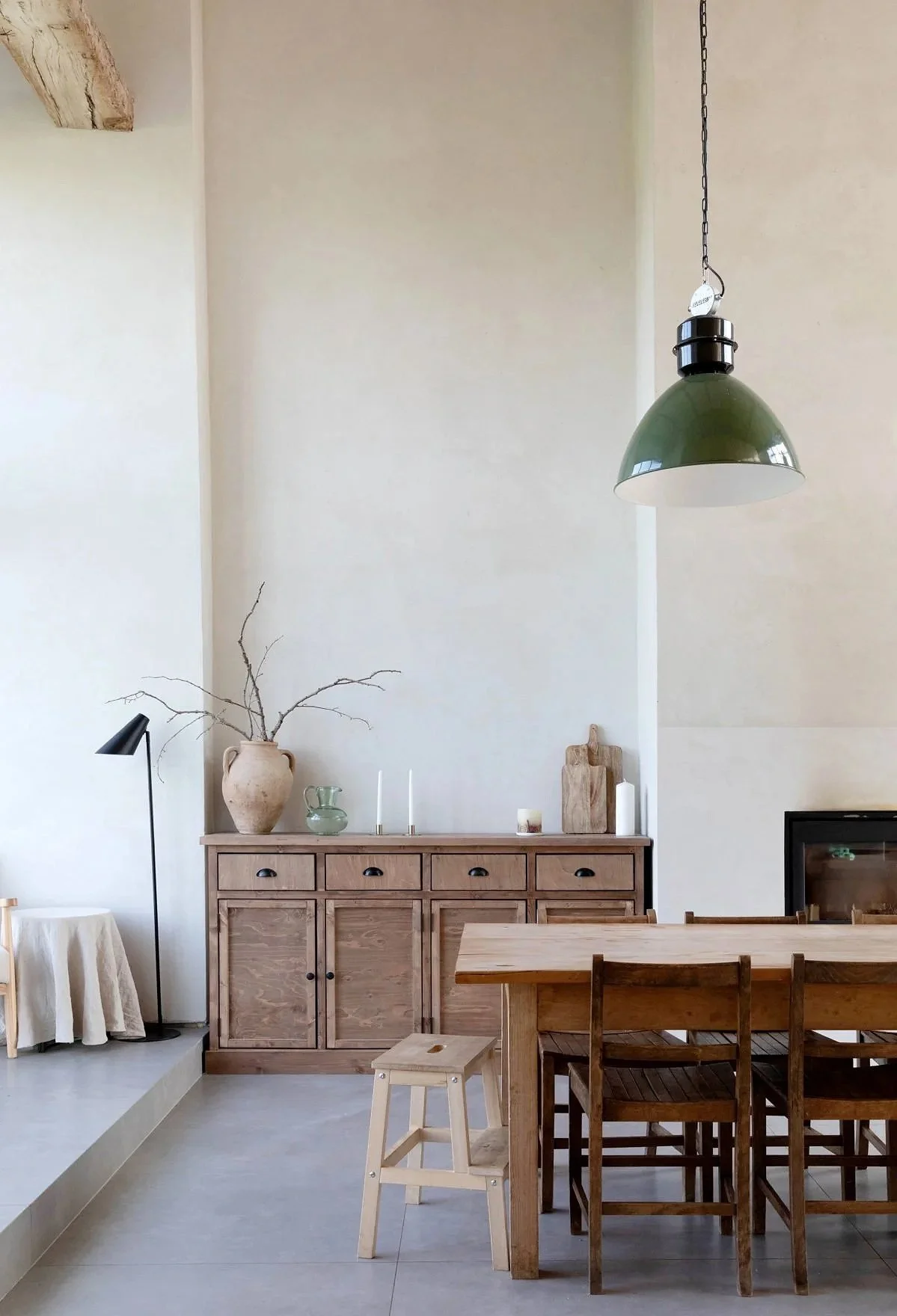 Interior of a minimalist dining room with a wooden dining table, four chairs, a wooden sideboard with decorative items including a vase with branches, candles, and small kitchen items, green pendant light, and neutral wall colors.