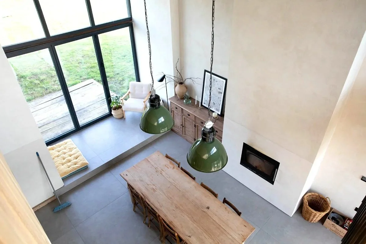 A high-angle view of a modern living dining area with a large wooden table, six chairs, two large green pendant lights, a built-in window seat with a cushion, a white armchair near large glass doors leading outside, a wooden sideboard with decorative