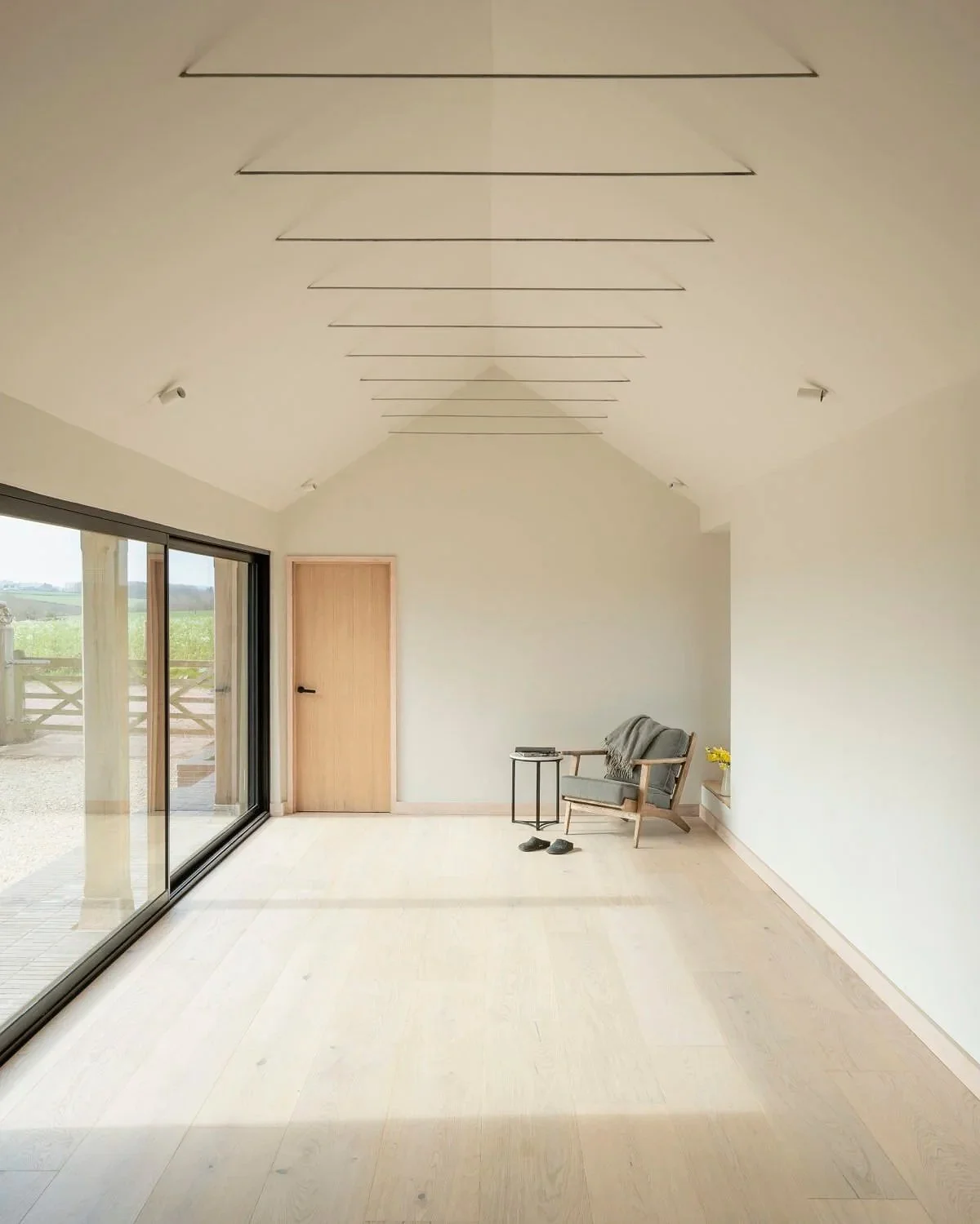 Minimalist living room with large sliding glass doors, a closed wooden door, a chair with a blanket, a small side table, and a pair of slippers on the light-colored wooden floor, with a view of a grassy outdoor area.