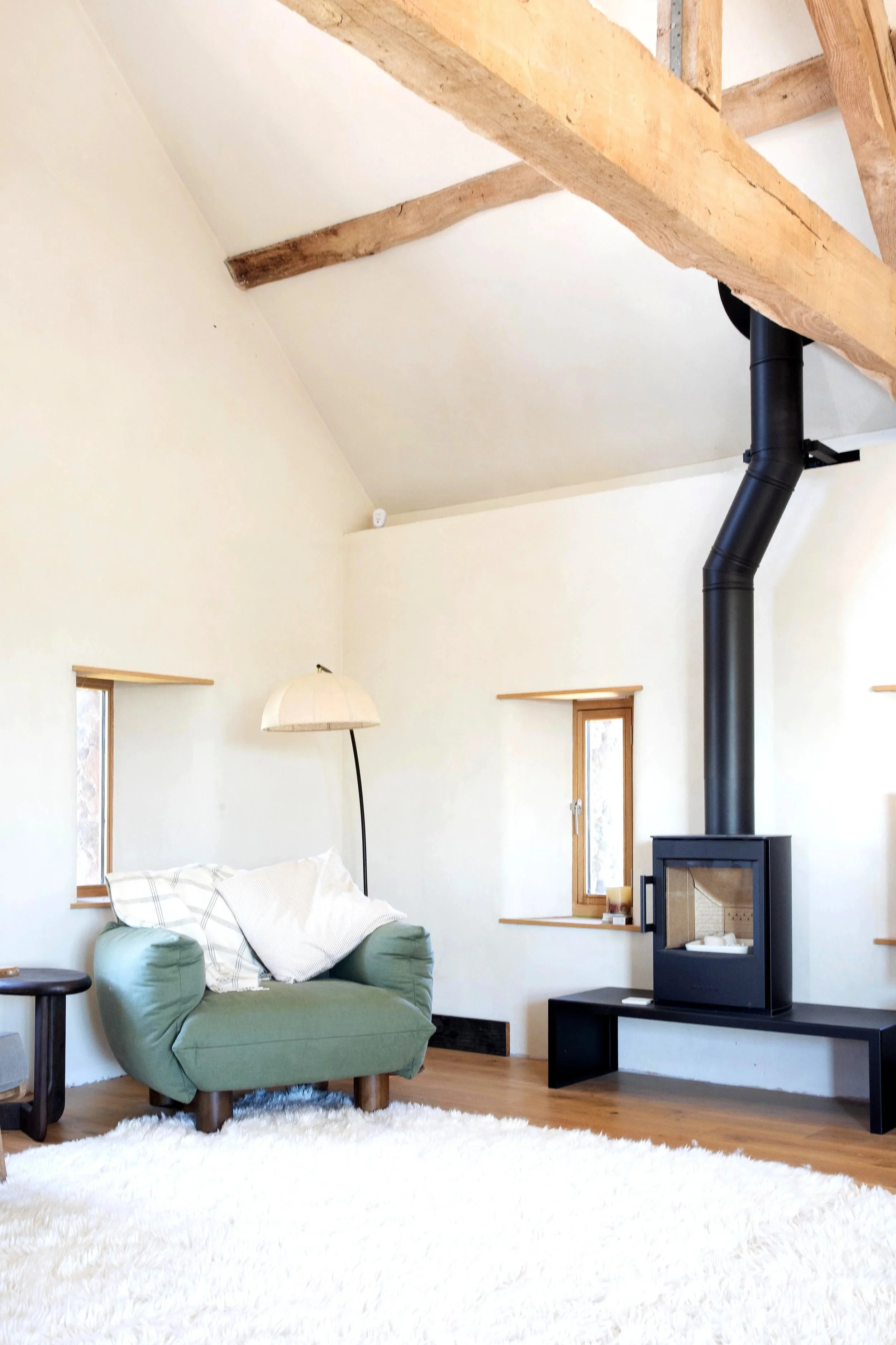 Living room with a green armchair, a white fluffy rug, a black stove with a chimney, and windows with wooden frames, in a room with white walls and wooden ceiling beams.
