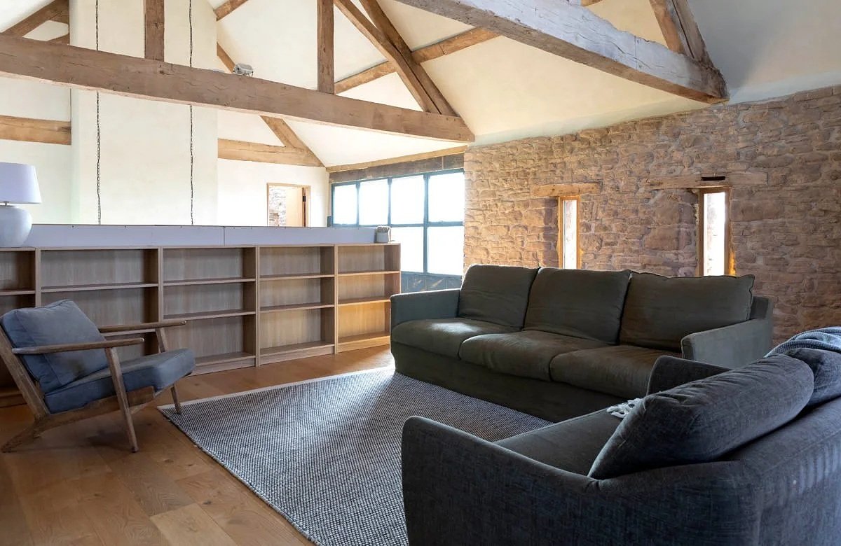 Living room with exposed wooden beams, brick wall, and natural light from windows. Contains two dark-colored sofas, an armchair, a rug, and a bookshelf.