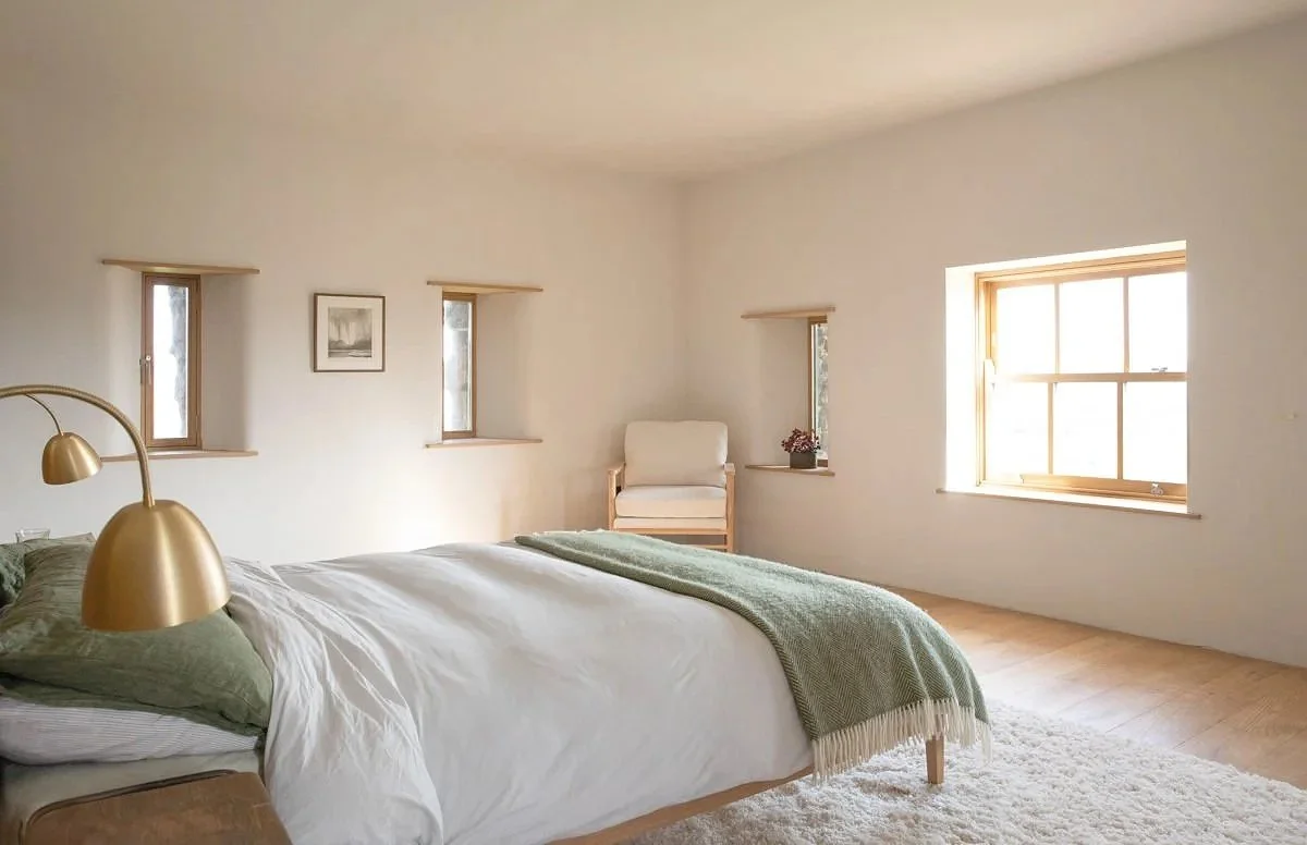 Bright bedroom with a large bed, beige armchair by the window, multiple small windows, light wood floors, and simple decor.