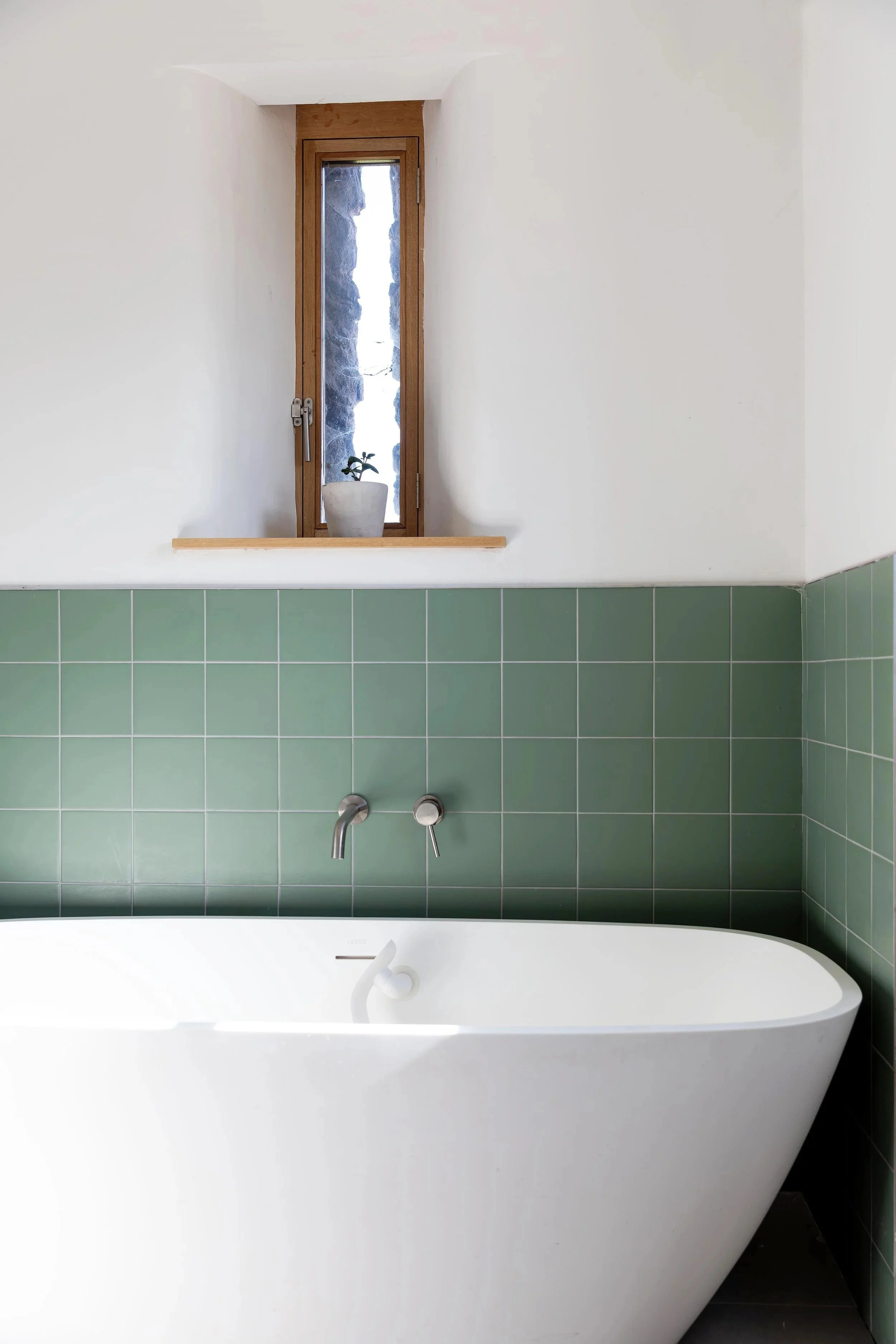 White bathtub in a bathroom with green tiled wall. Above the tub, a ledge with a small plant in a white pot. Behind the plant is a small window with a wooden frame, through which natural light and rocks are visible.