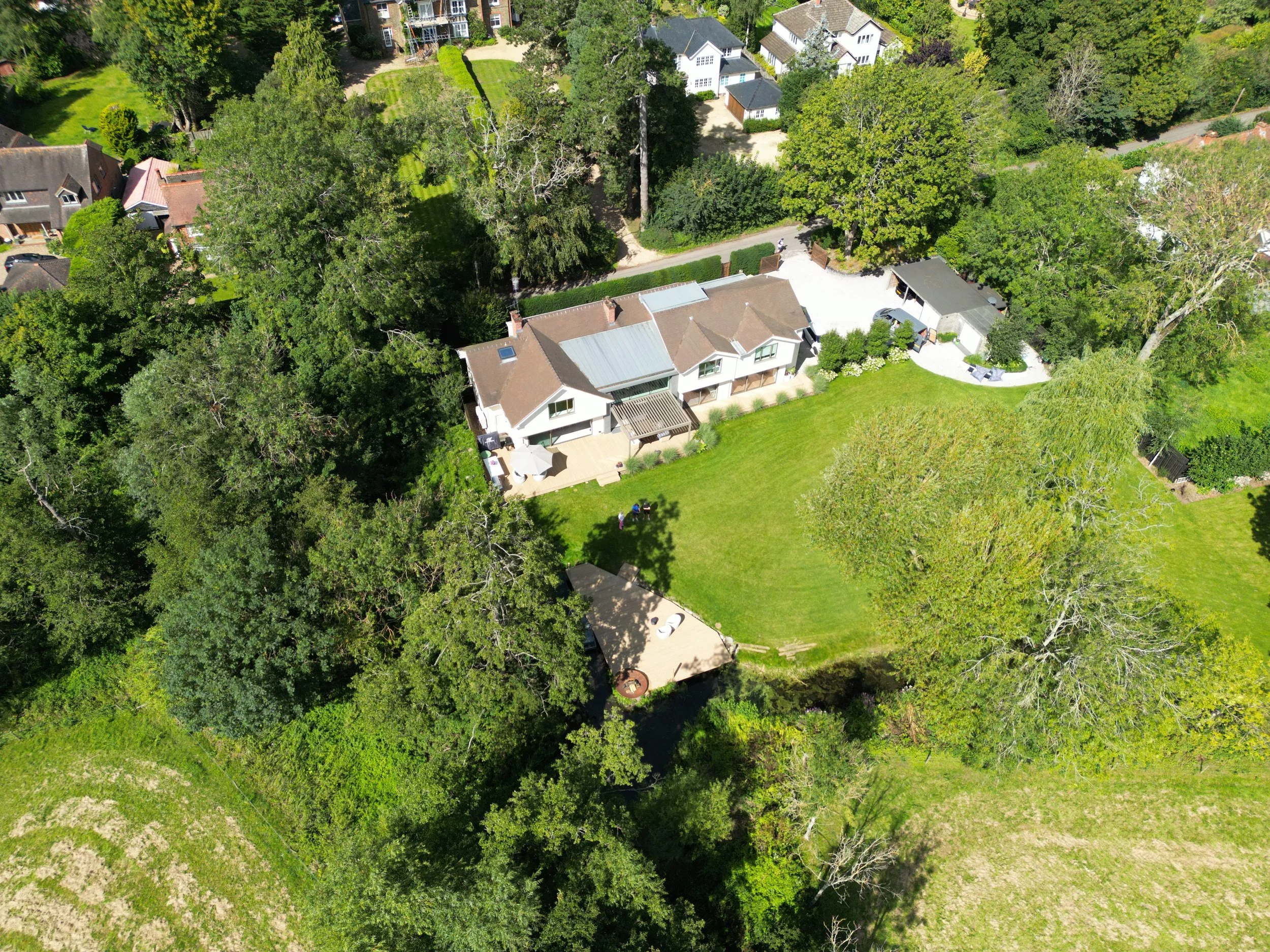 Aerial view of a residential backyard with a lawn, trees, a patio area, and surrounding houses.