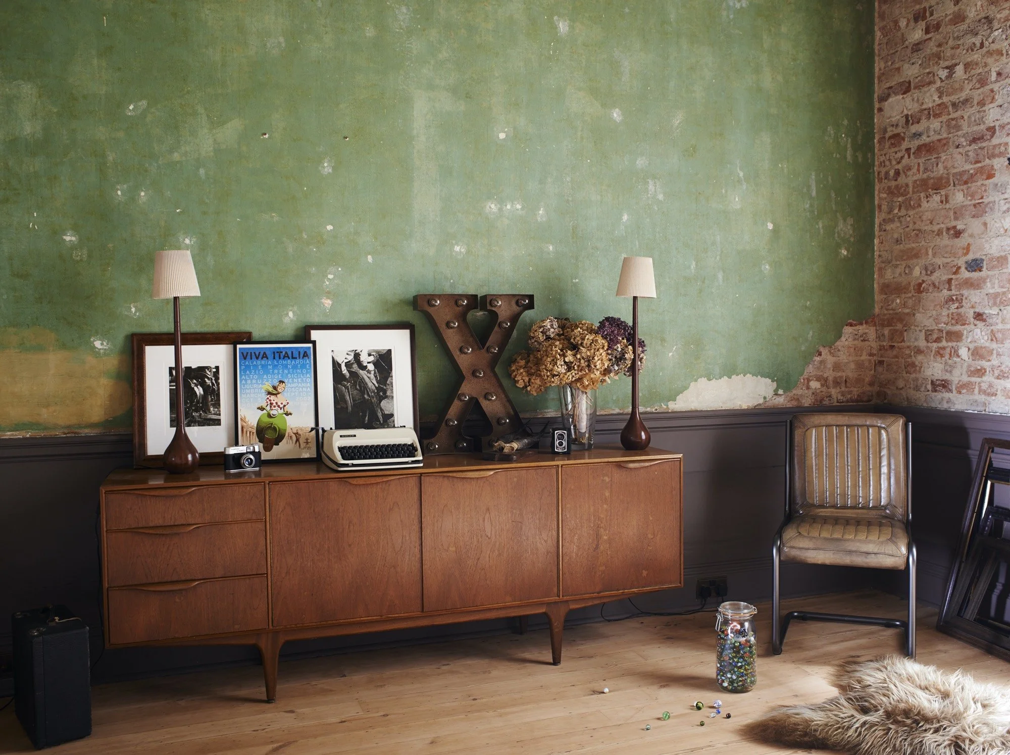 Vintage wooden sideboard against a distressed green wall with an exposed brick corner. Decor includes framed photos, a large letter 'X' with light bulbs, a vase with dried flowers, two table lamps, a camera, typewriter, and a small camera. A leather 