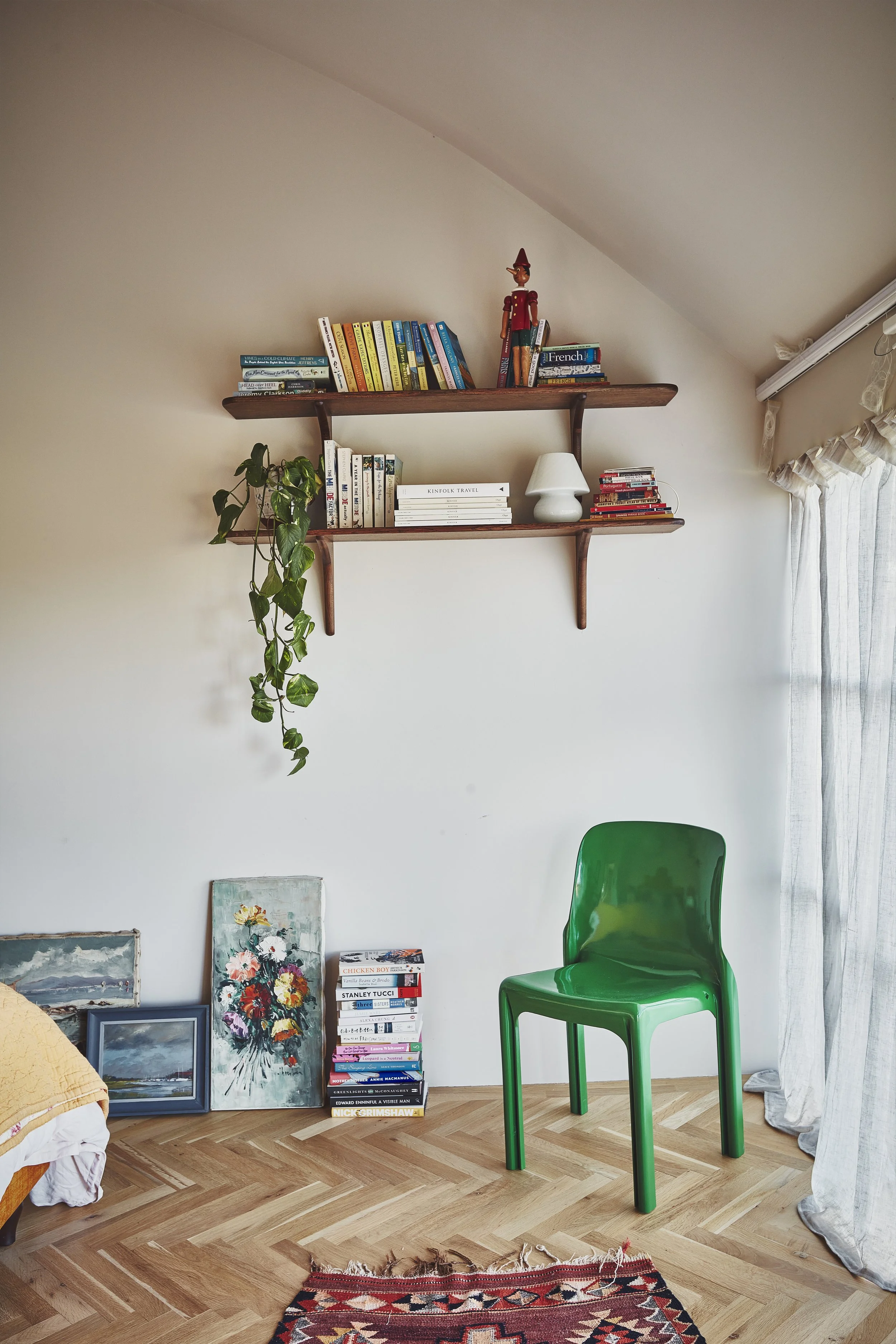 A room with a wooden parquet floor, a green plastic chair, a stack of books, paintings leaning against the wall, a white lamp, a red and black rug, and two floating wooden shelves with books and a wooden Pinocchio puppet, next to a window with white 