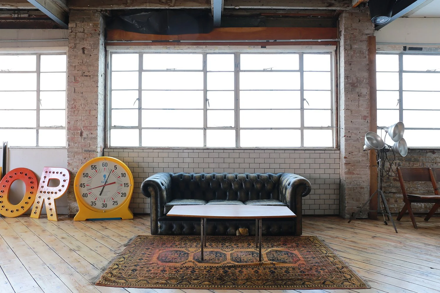 Interior of a stylish loft with large industrial windows, a black leather tufted sofa, a wooden coffee table, a vintage patterned rug, a large yellow clock, decorative letter signs, and studio lighting on a tripod.