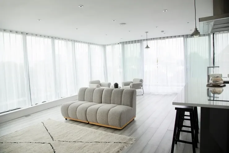 Bright, modern living room with large windows, white curtains, a plush gray sofa, two white armchairs, a white rug, and a white kitchen island with black stools.