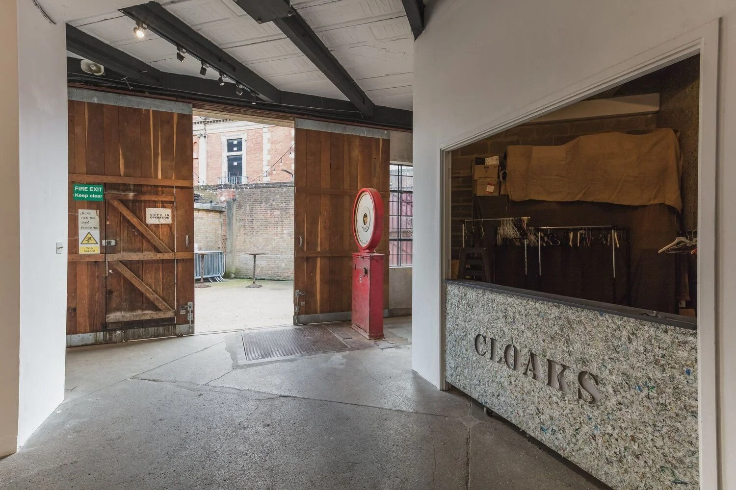 Interior of a shop with a granite counter labeled 'CLOAKS', an open wooden barn door leading outside, a red vintage scale, and a fire exit door.