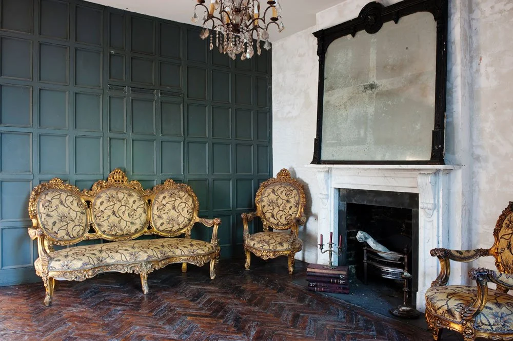 Living room with green paneled wall, ornate gold and floral upholstered furniture, a white fireplace with a large mirror above, and a chandelier hanging from the ceiling.