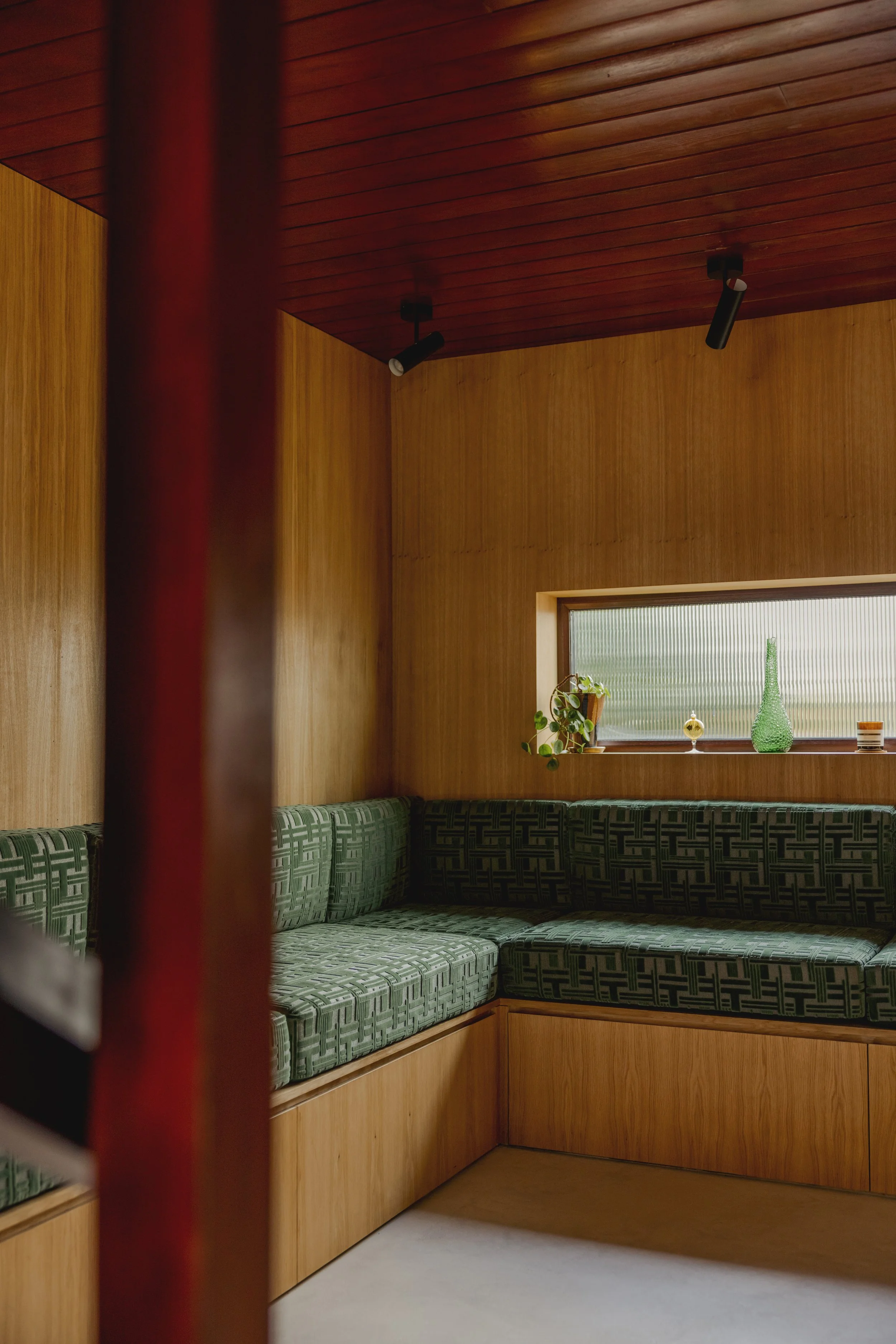 Corner of a room with wooden walls and ceiling, featuring a large window with decorative vases and a potted plant. A built-in green patterned upholstered bench.'