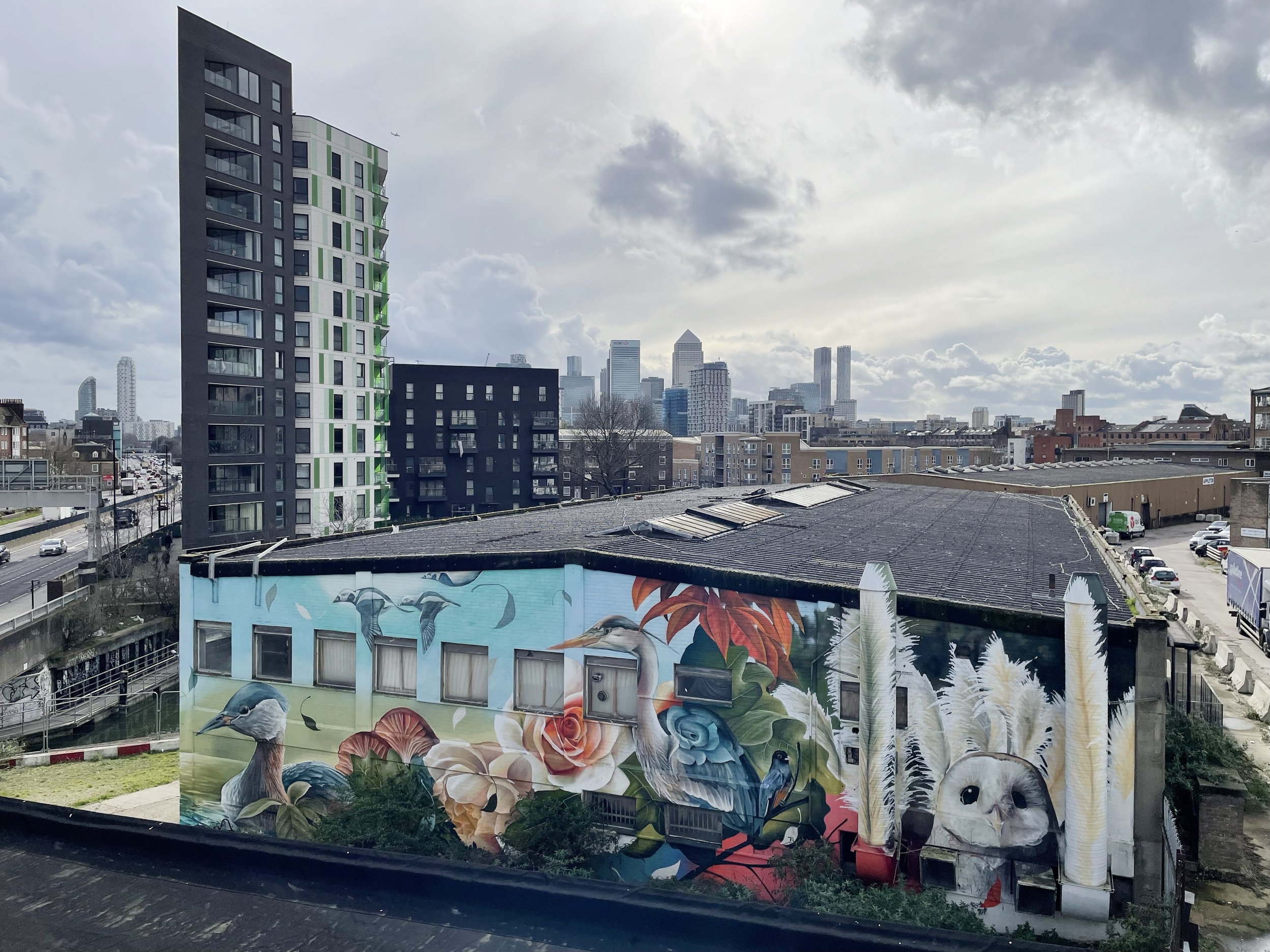 City skyline with skyscrapers, a mural featuring birds and flowers on a building, and cloudy sky