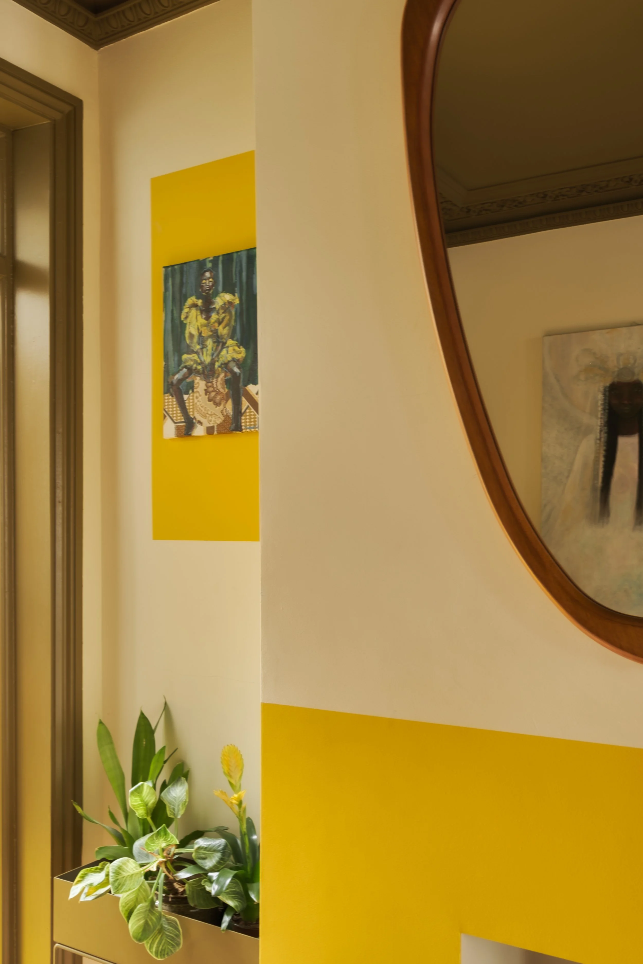 Interior view of a room showing a mirror with a wooden frame, a yellow and white painted wall, a plant in a black pot, and two paintings on the wall, one with a person wearing yellow and the other with a person in white.